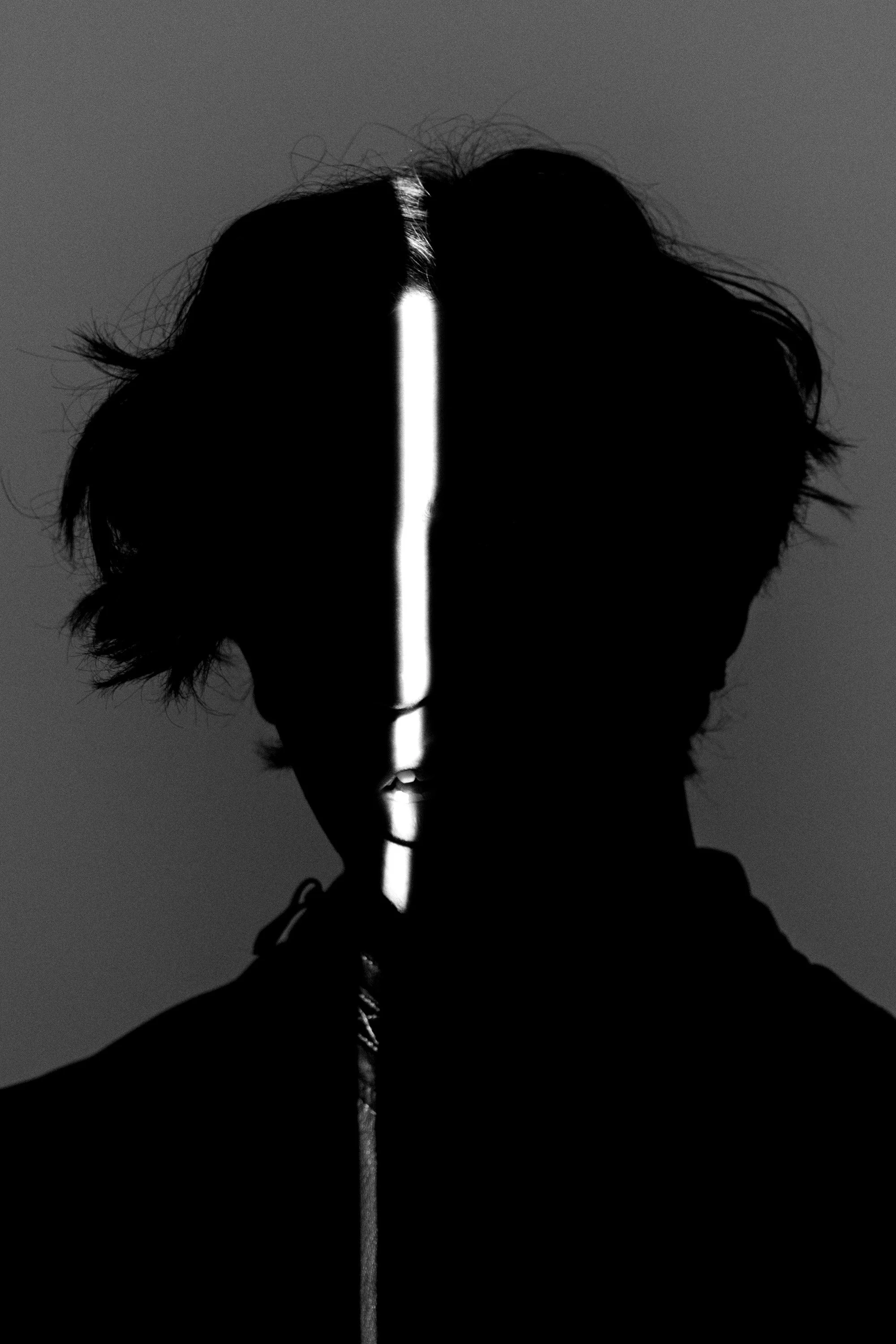 Silhouetted person with disheveled hair facing slightly right, with light reflecting off the face creating a vertical line down the center, against a dark background.