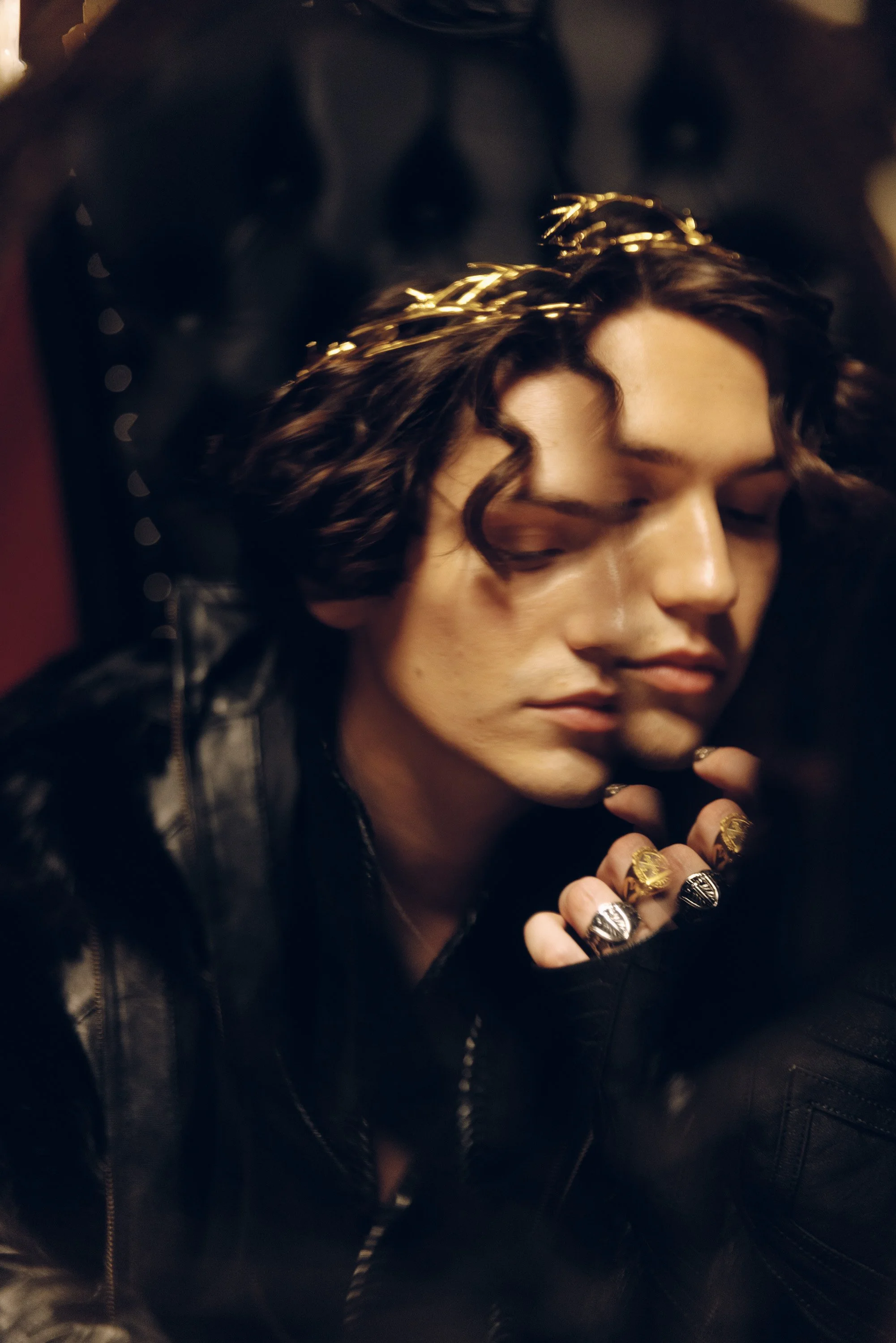 Close-up of a young woman with curly brown hair and a gold crown, wearing a black leather jacket, with rings on her fingers, resting her hand near her face.