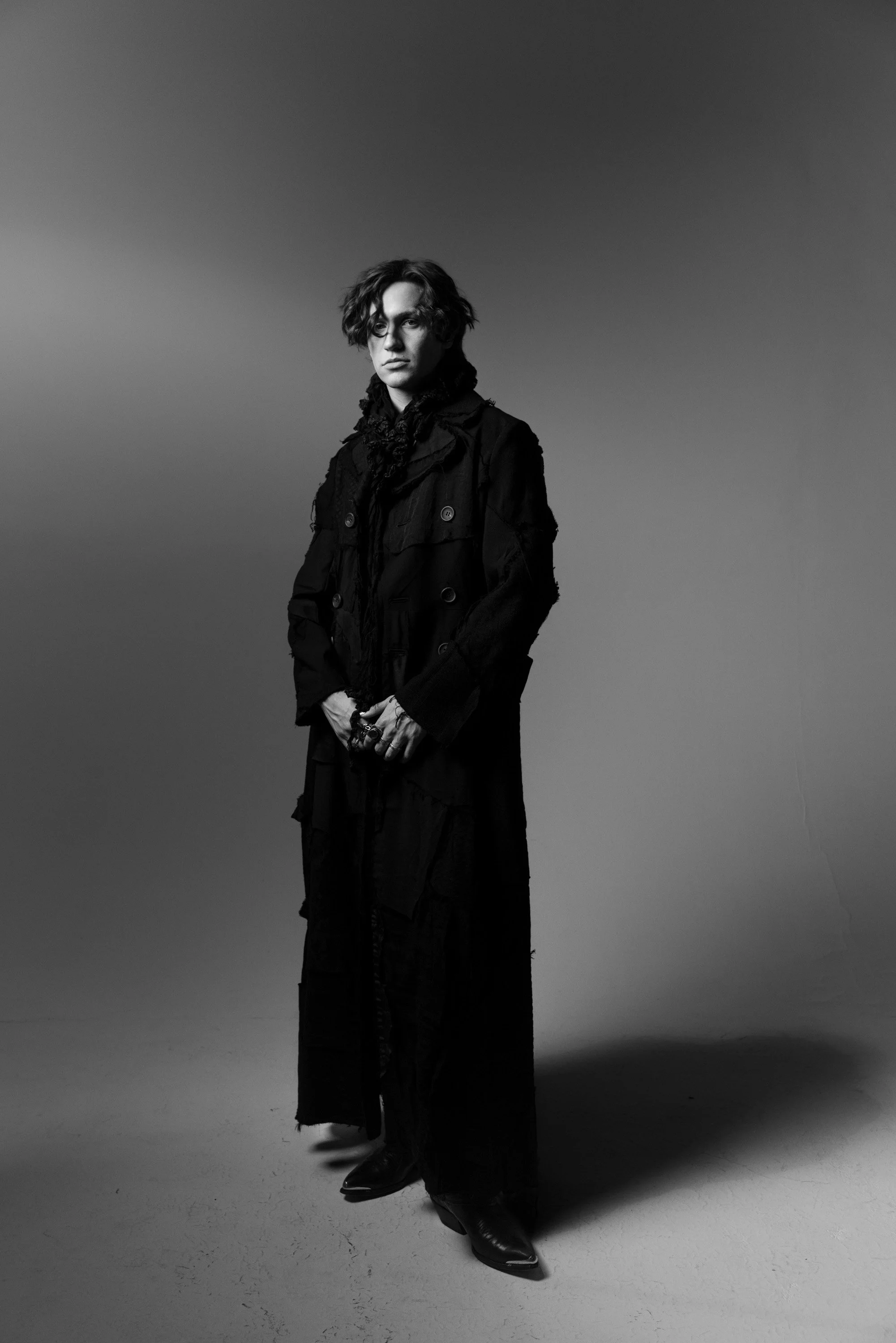 A person dressed in dark clothing, wearing boots, standing against a plain backdrop in a photo with shadows on the floor.