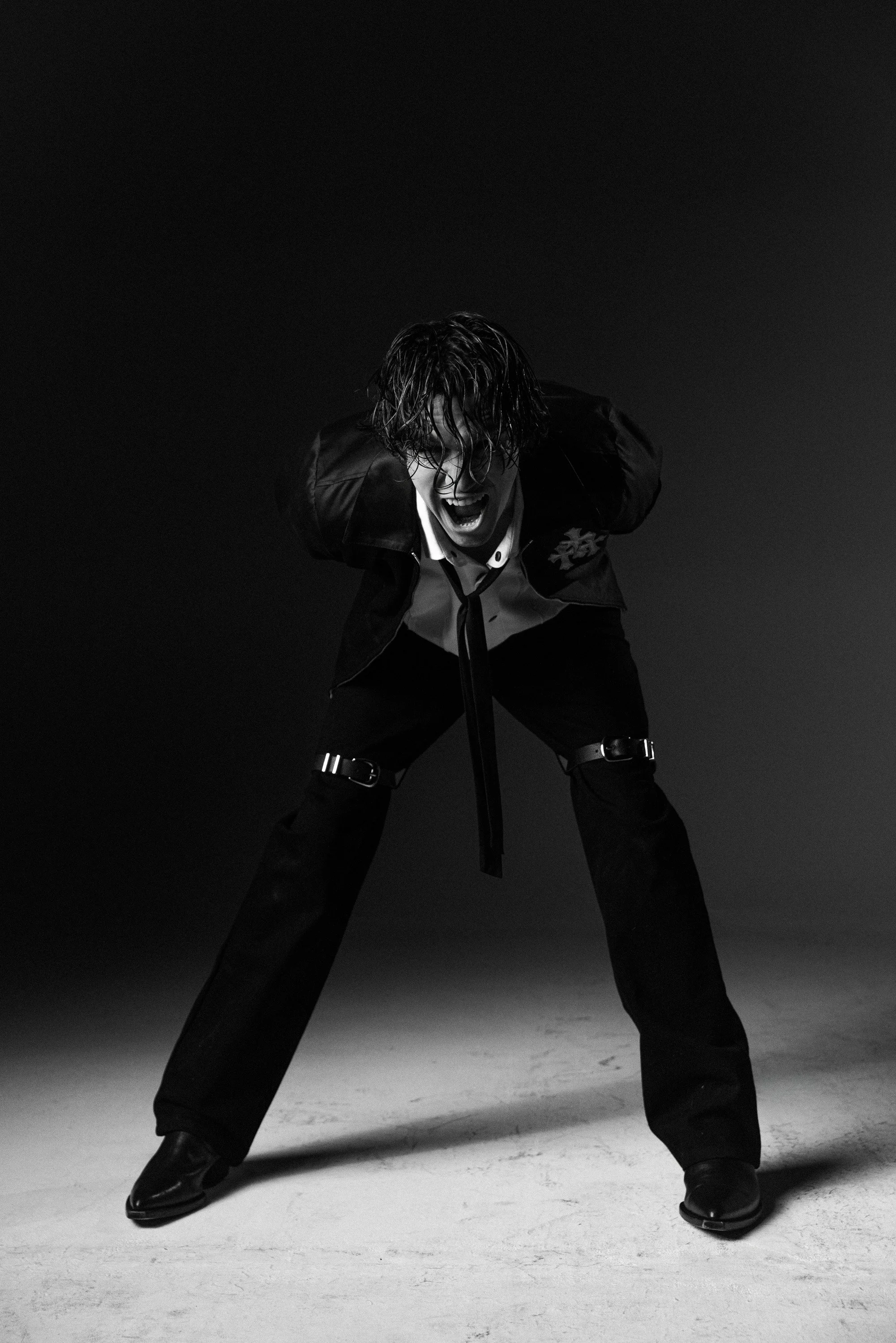A person with wet hair, wearing a leather jacket and black pants, stands in a wide stance with legs apart and arms behind their back, looking down with an intense expression in a dark studio setting.