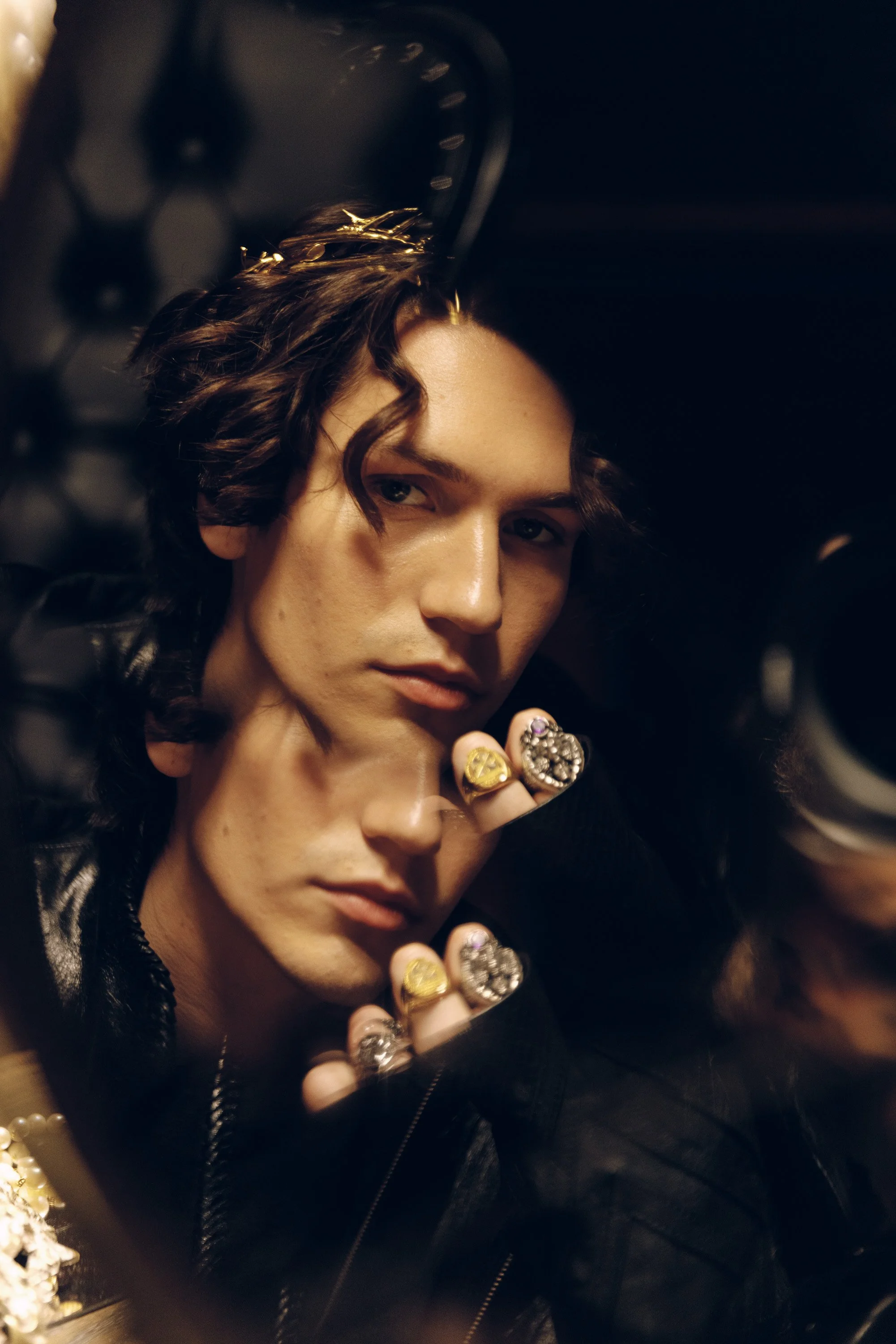 A person with long, wavy brown hair wearing multiple rings, a pearl bracelet, and a black leather jacket, with a dark, padded background.