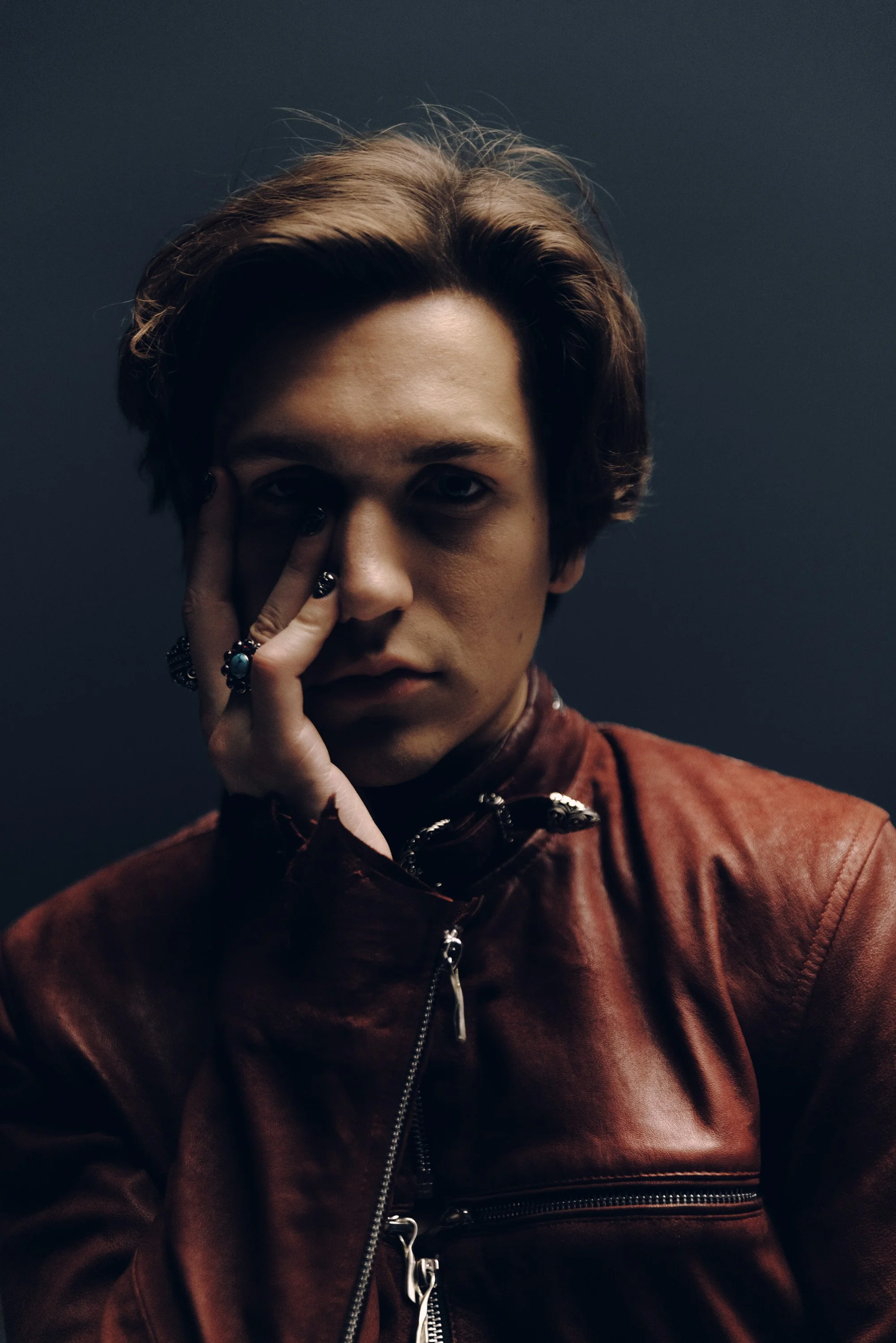 A young person with medium-length brown hair and light skin, wearing a reddish-brown leather jacket and multiple rings, partially covering their face with their hand, against a dark gray background.