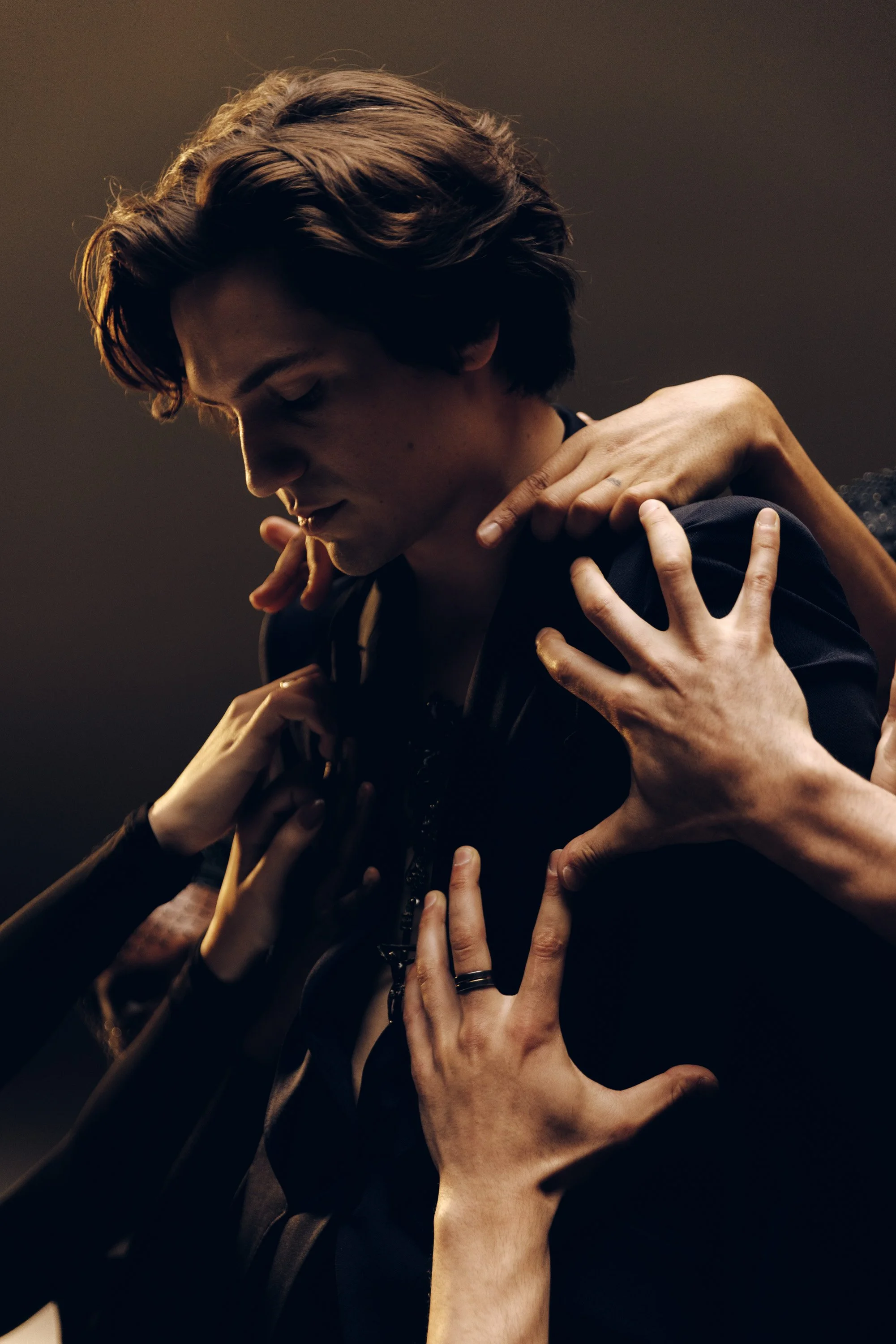 Multiple hands reaching towards and touching a young man's shoulders, neck, and upper chest from various angles, in a dark setting.