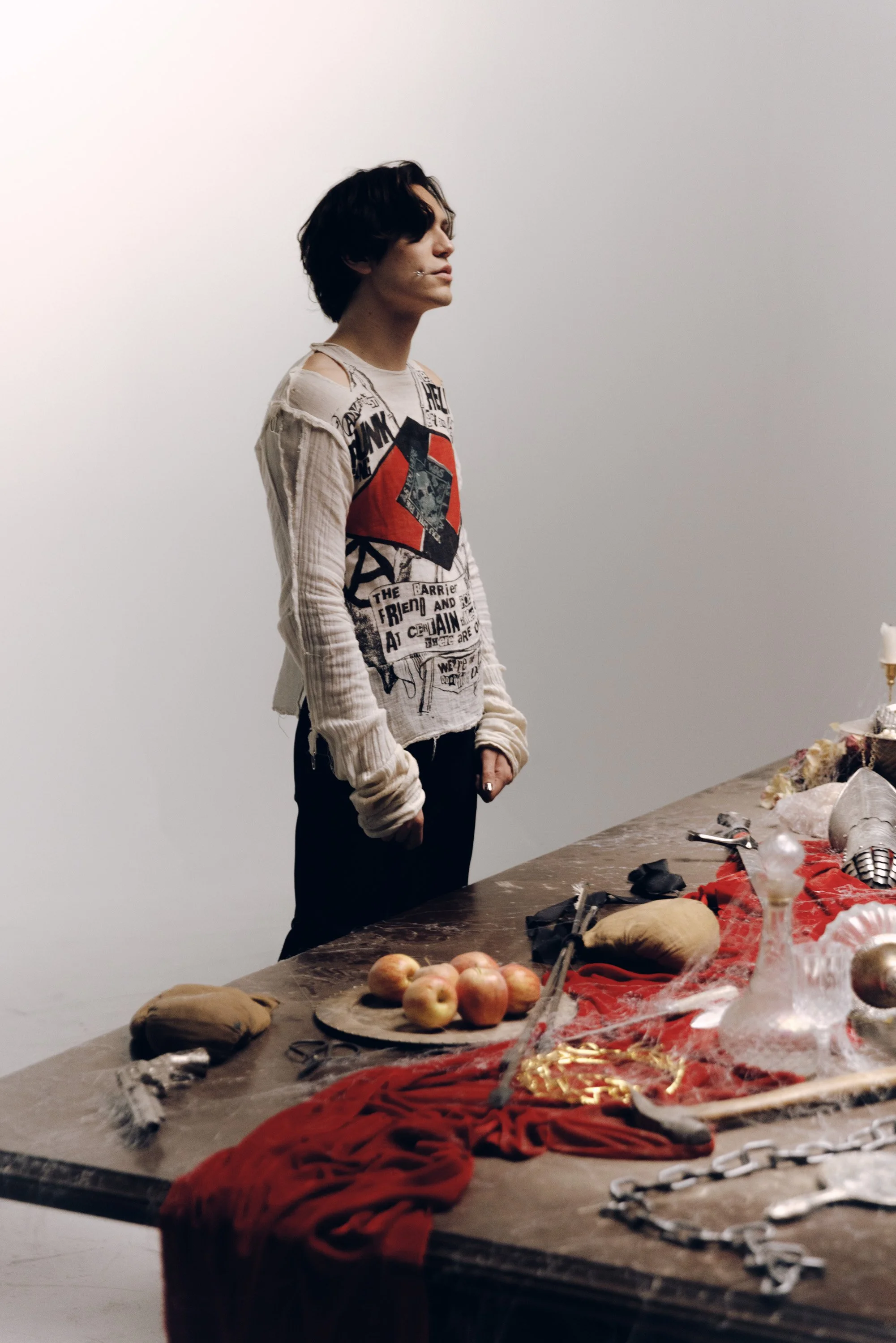 A person with dark, wavy hair stands by a table covered with various items, including bread, apples, jewelry, and fabric. The person is wearing a long-sleeved shirt with graphic text and images, and has a contemplative expression. The background is p