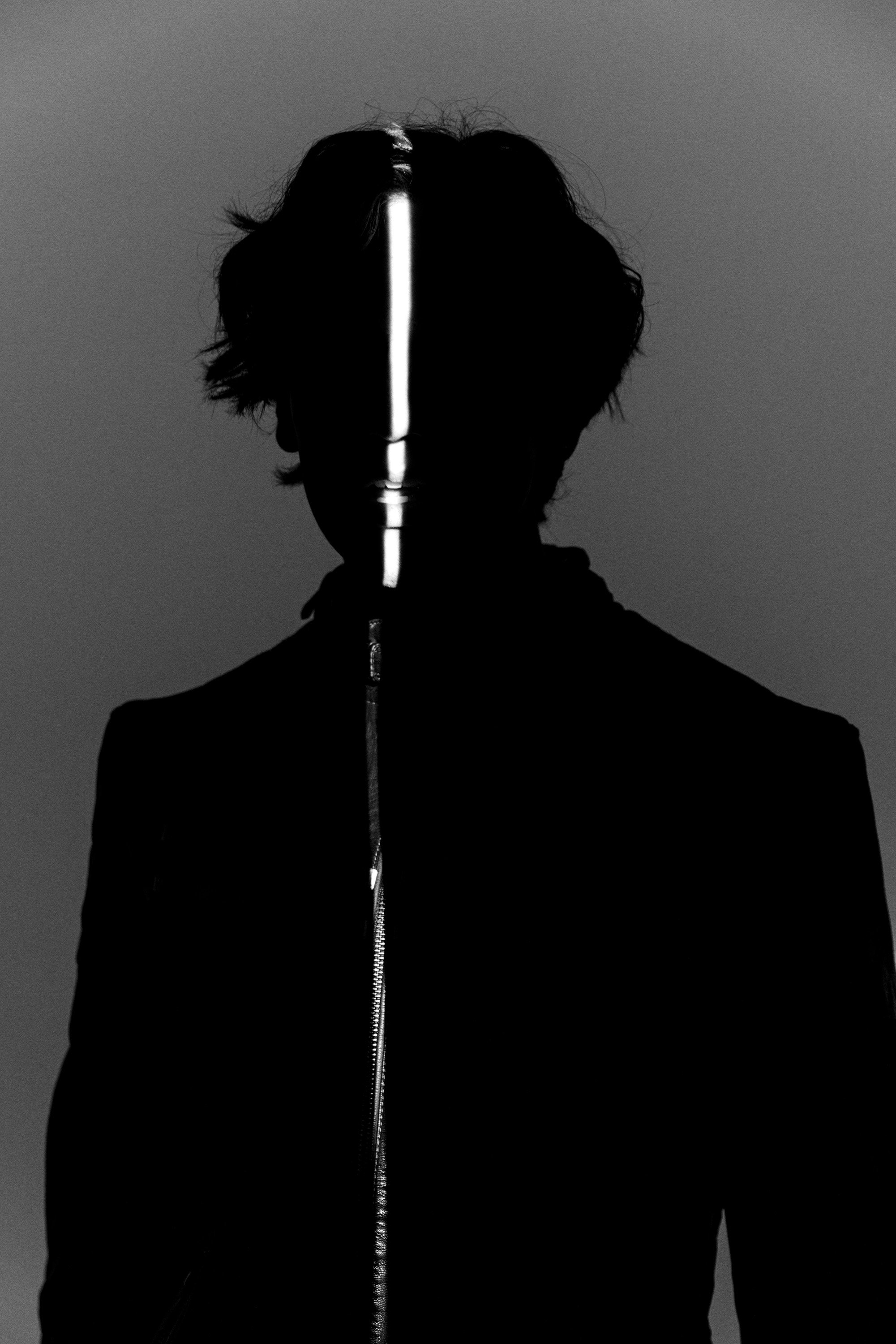 A person with dark, messy hair standing against a gray background, wearing a black zip-up jacket with a reflective strip along the zipper.