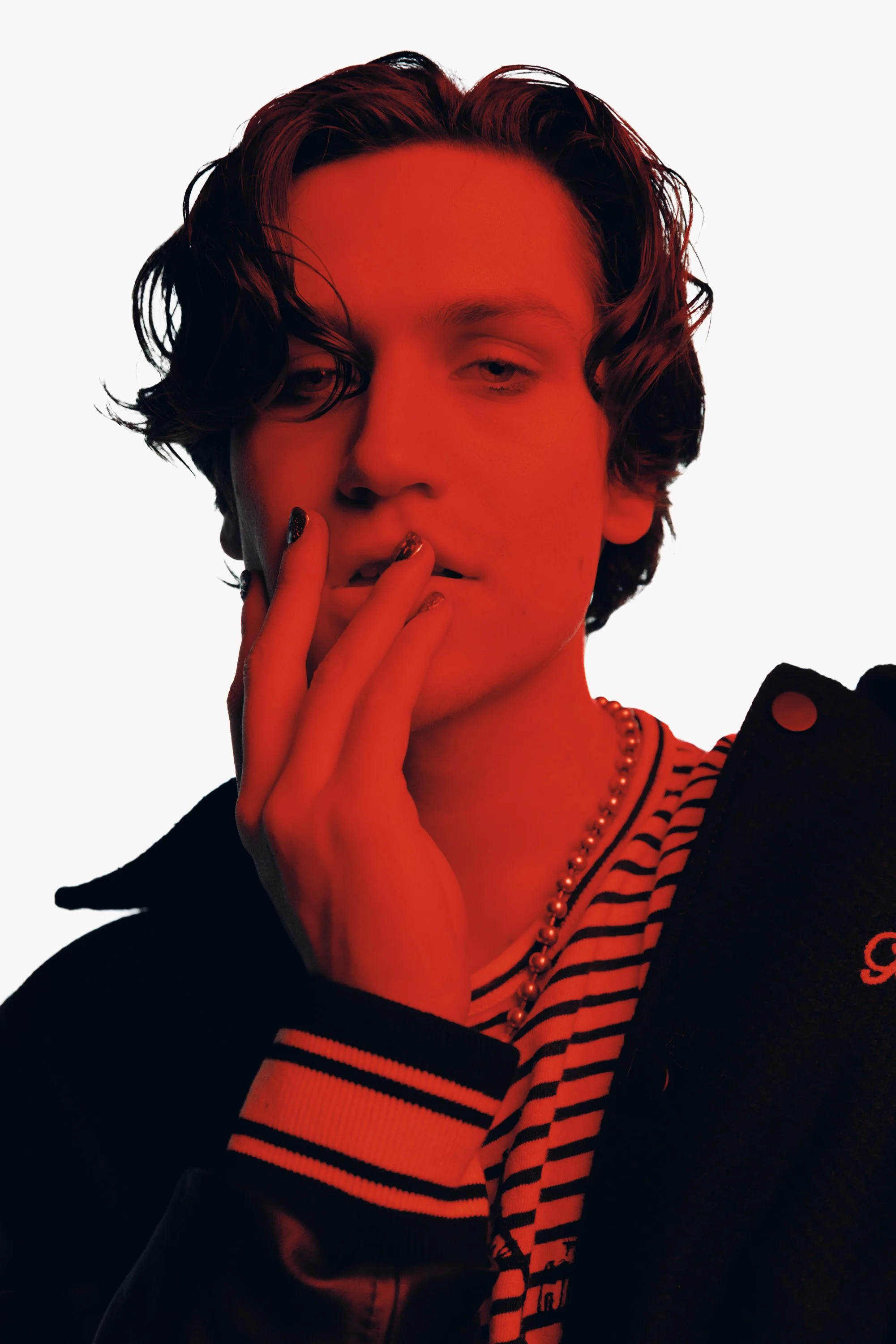 A person with dark wavy hair and makeup, wearing a striped shirt, a beaded necklace, and a dark jacket, striking a contemplative pose with fingers near their lips against a plain white background.