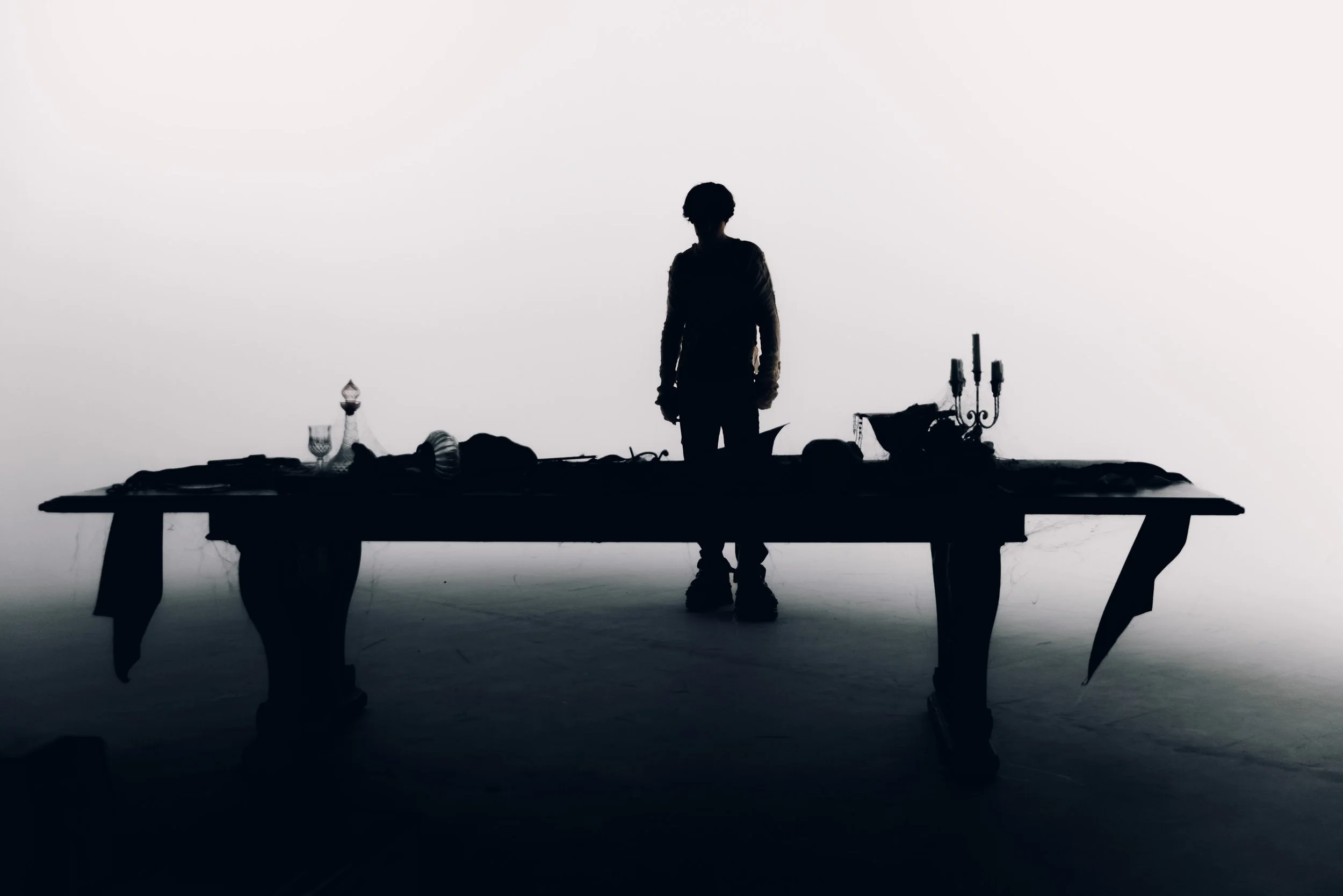 Silhouette of a person standing behind a table with various glassware and decorative objects, viewed in profile against a plain, bright background.