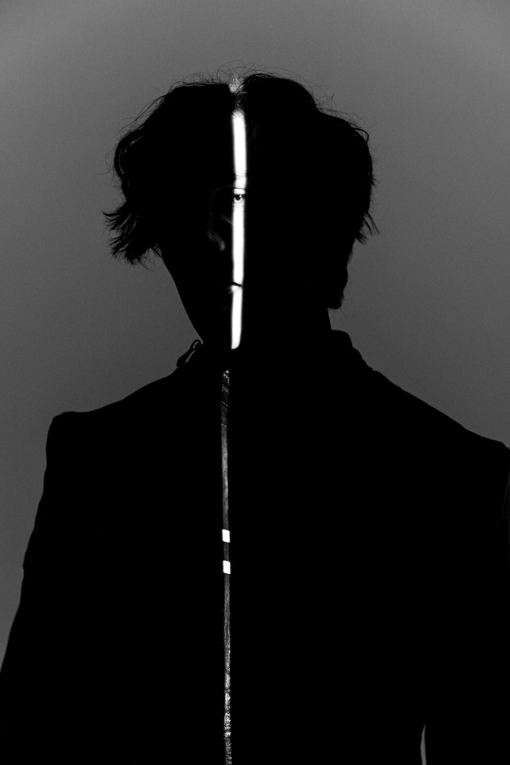 Black and white portrait of a person with dark, wavy hair, with a vertical glowing line running from top to bottom through the center of the face, obscuring one eye and part of the face.