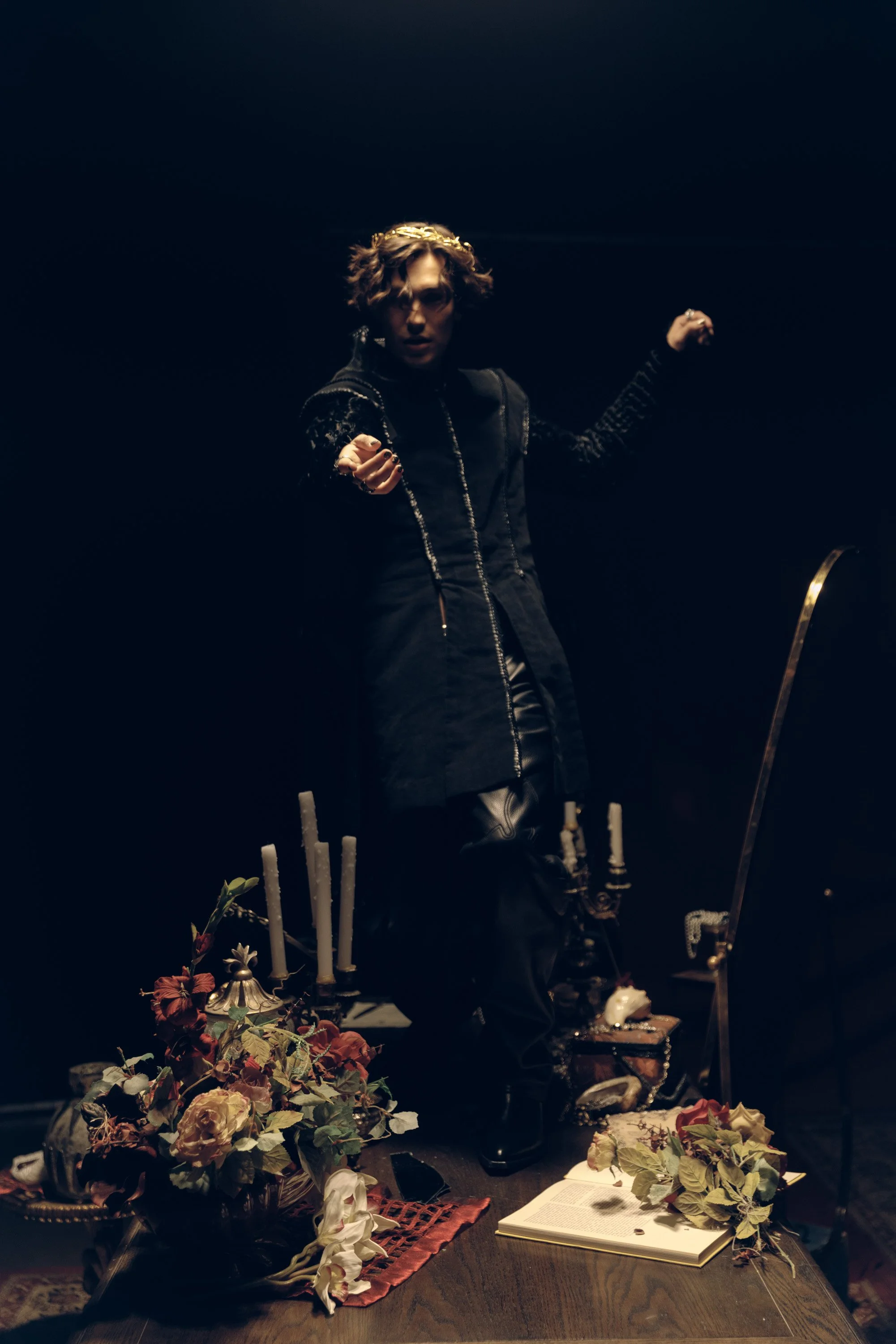 A woman with short wavy hair wearing dark clothing, standing with arms raised, in a dimly lit scene with books, flowers, candles, and religious items.