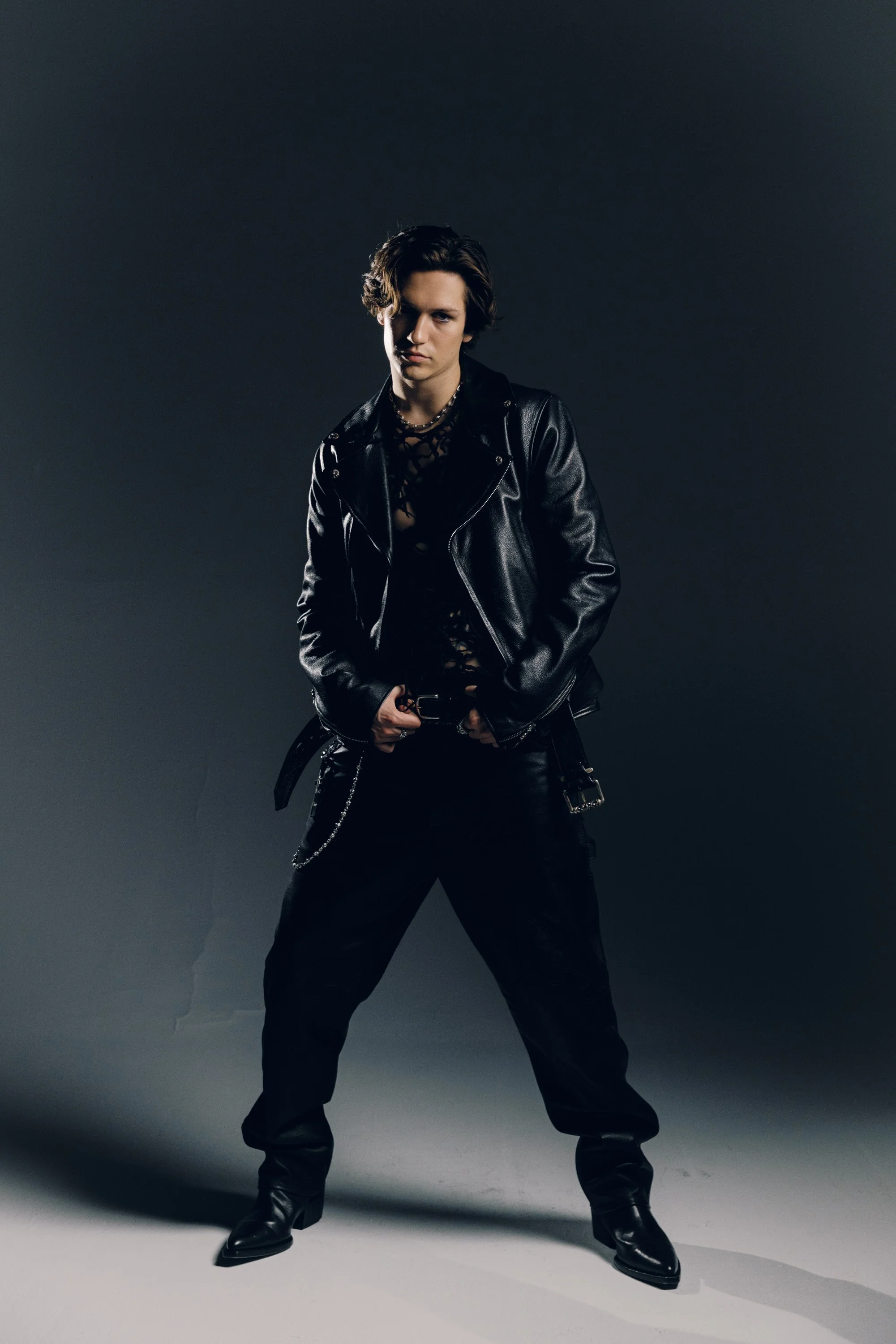 Young person with medium length dark hair wearing a black leather jacket, black pants, and boots standing against a dark gradient background.