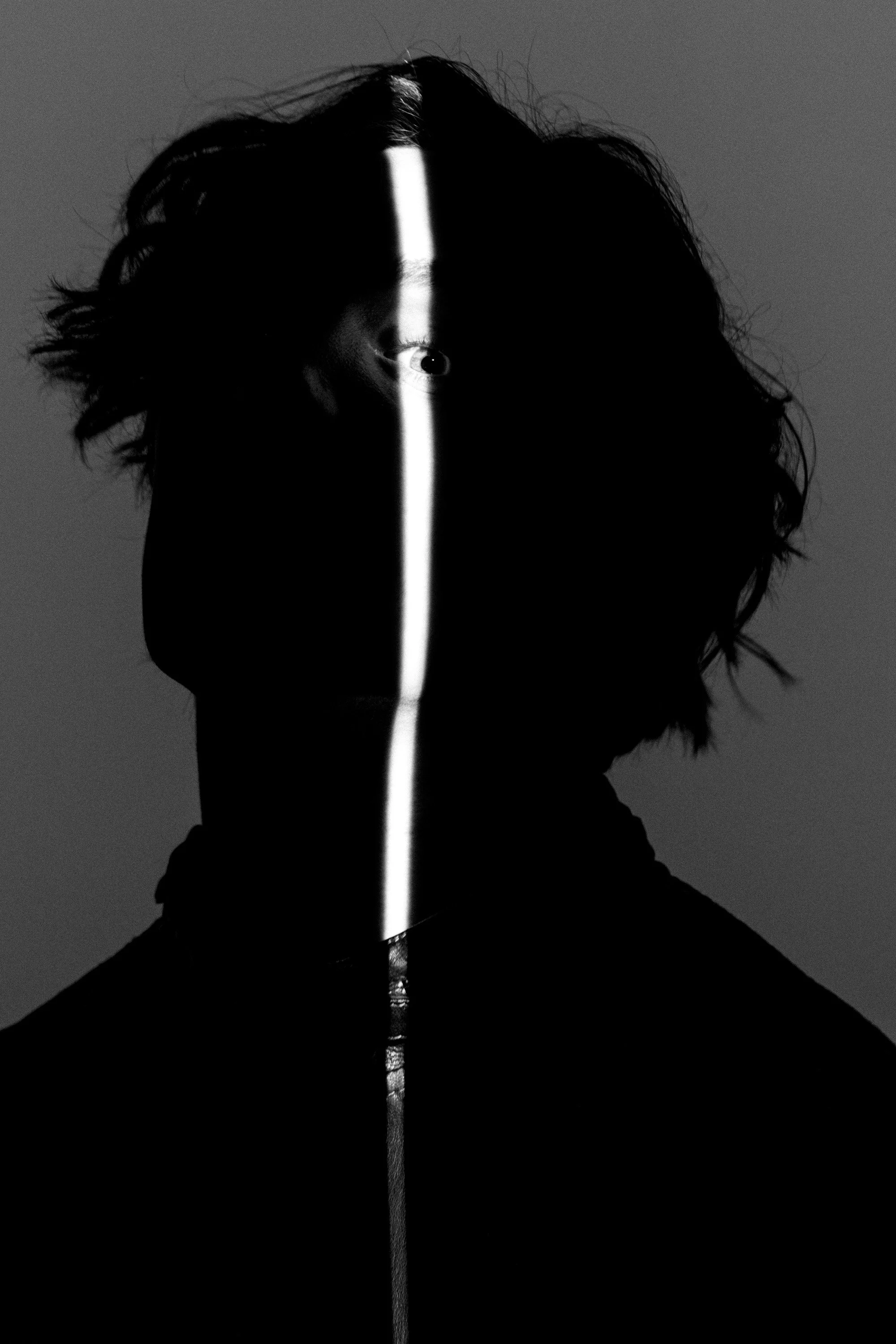 Black and white photo of a person's face with half illuminated and half in shadow, a vertical illuminated line running down the center of the face, revealing one eye and part of the nose.