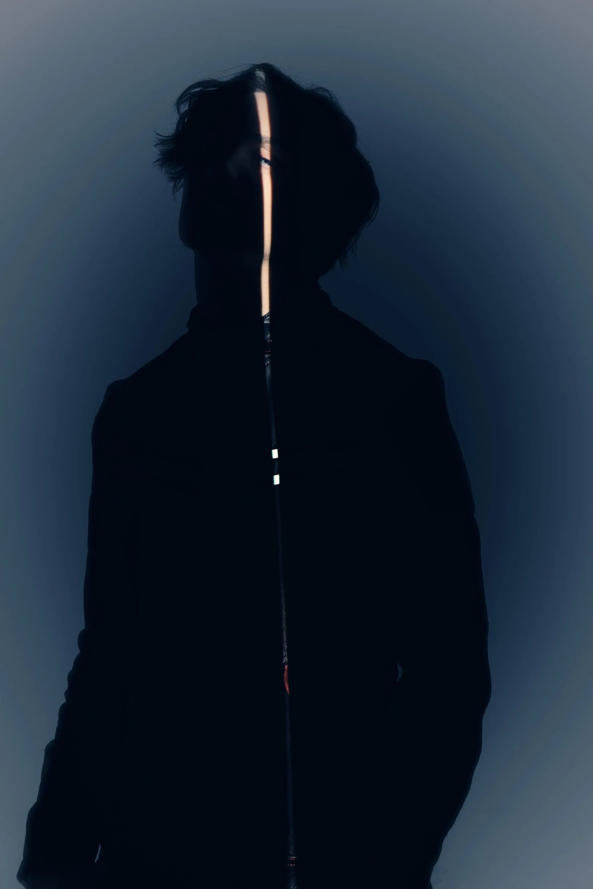 Silhouette of a person with a vertical light streak running down the face, messy hair, wearing a dark jacket, with a dark background.