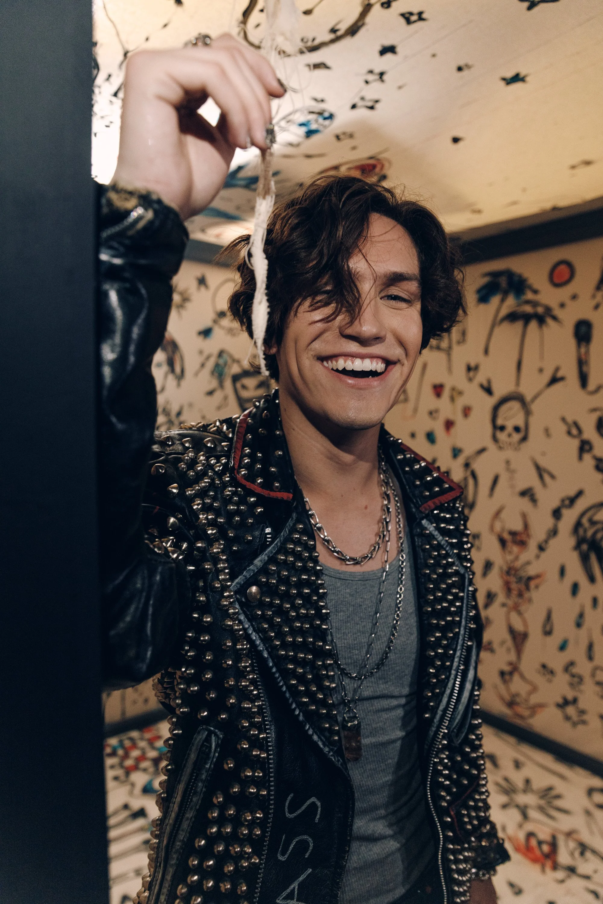 A young man with dark, curly hair smiling while holding a small object or charm with strings in his hand, wearing a spiked leather jacket and layered necklaces, standing in an art studio or gallery with a wall decorated with various drawings, sketche