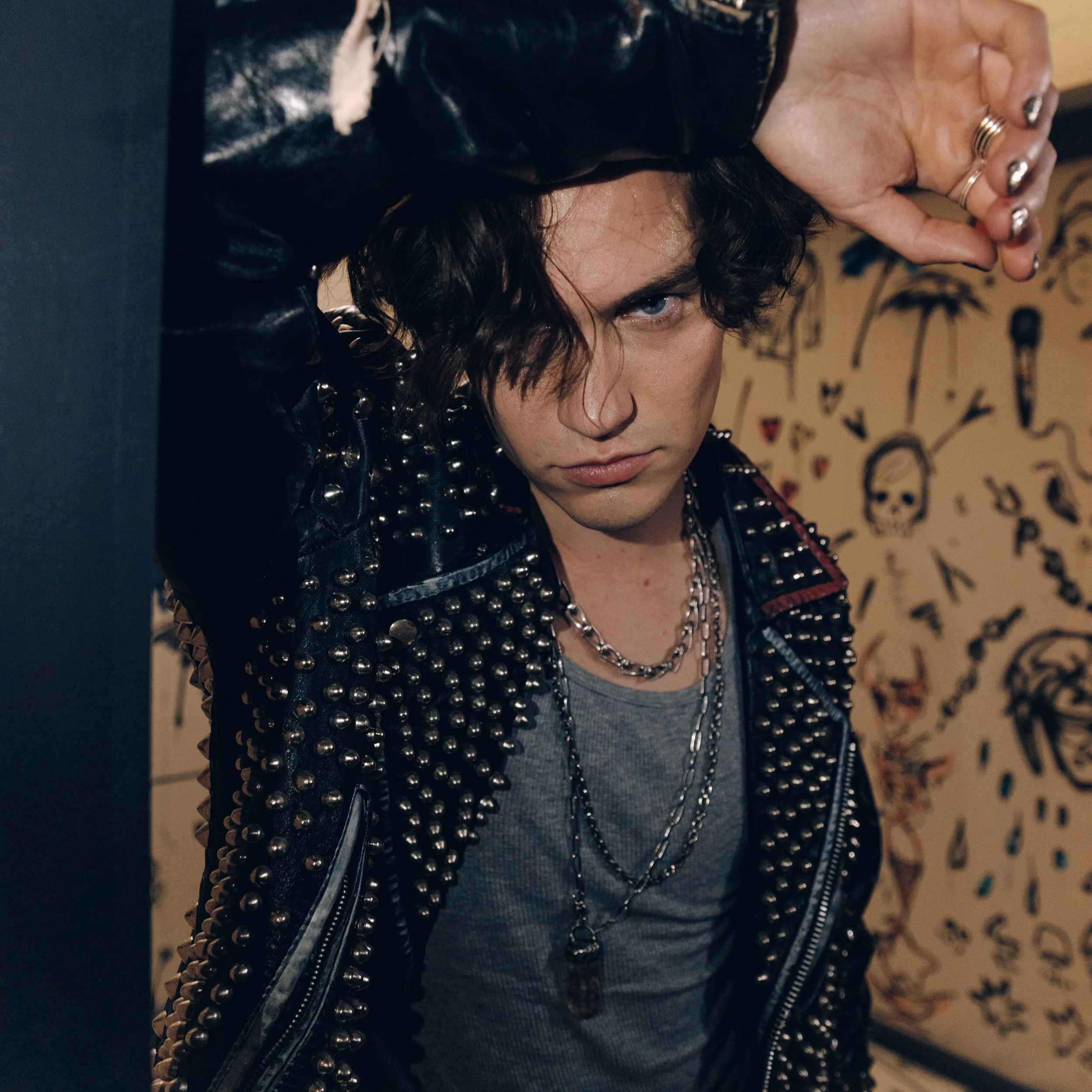 Close-up of a person with curly dark hair wearing a black leather jacket with metal studs, a gray t-shirt, and layered silver necklaces, holding one hand up to their face with metallic manicure, partially covering of the face.