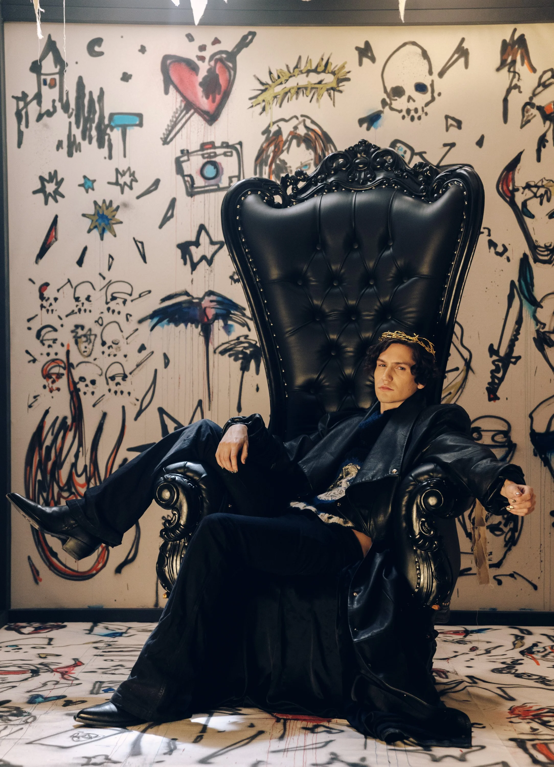 Young man sitting on a large, ornate black leather throne with a high backrest, in front of a graffiti-style wall featuring colorful drawings of skulls, hearts, stars, a camera, and abstract shapes.