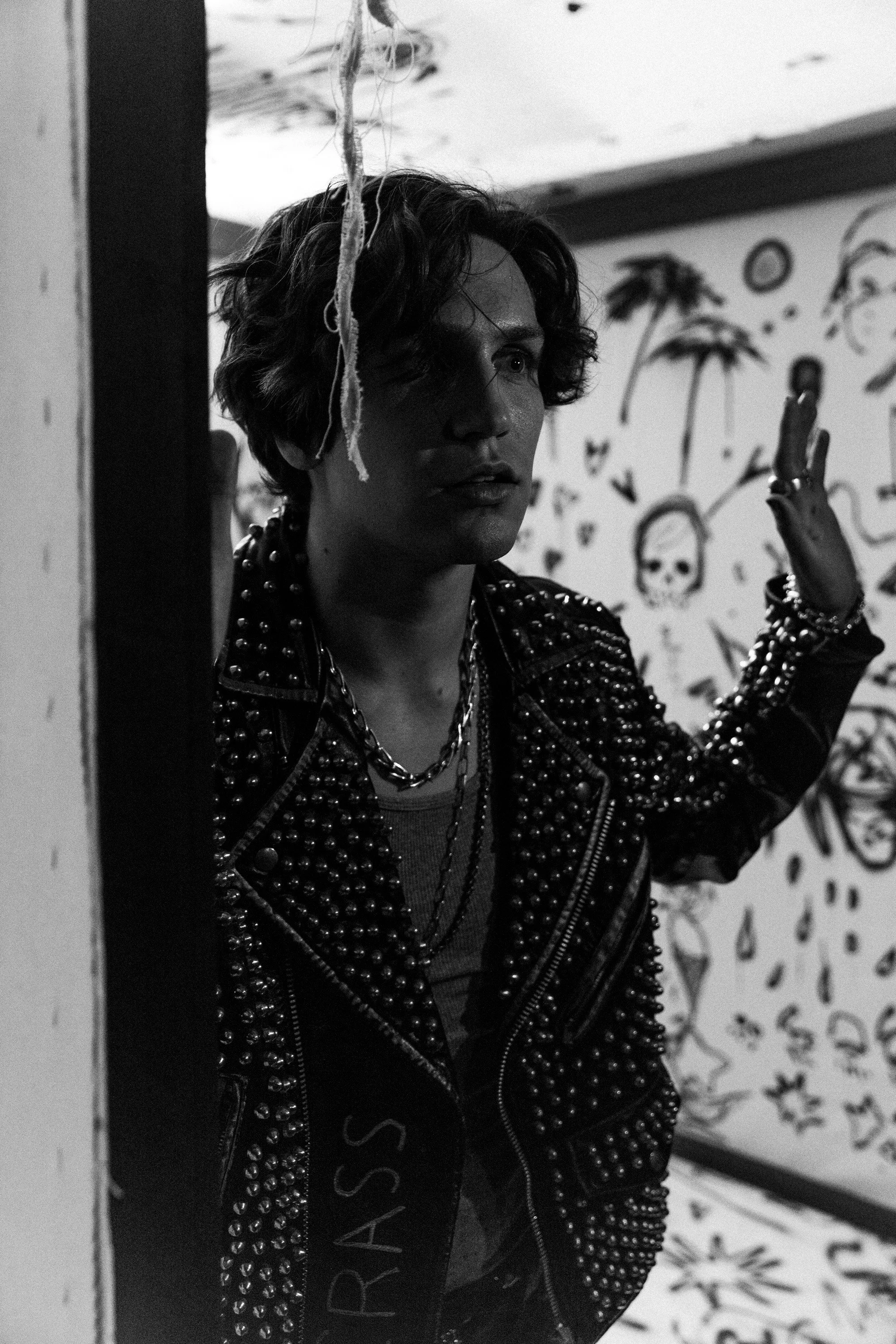 Black and white photo of a young person with curly hair, wearing a studded leather jacket with chains, standing against a wall with graffiti art of palm trees and skulls, raising their hand.