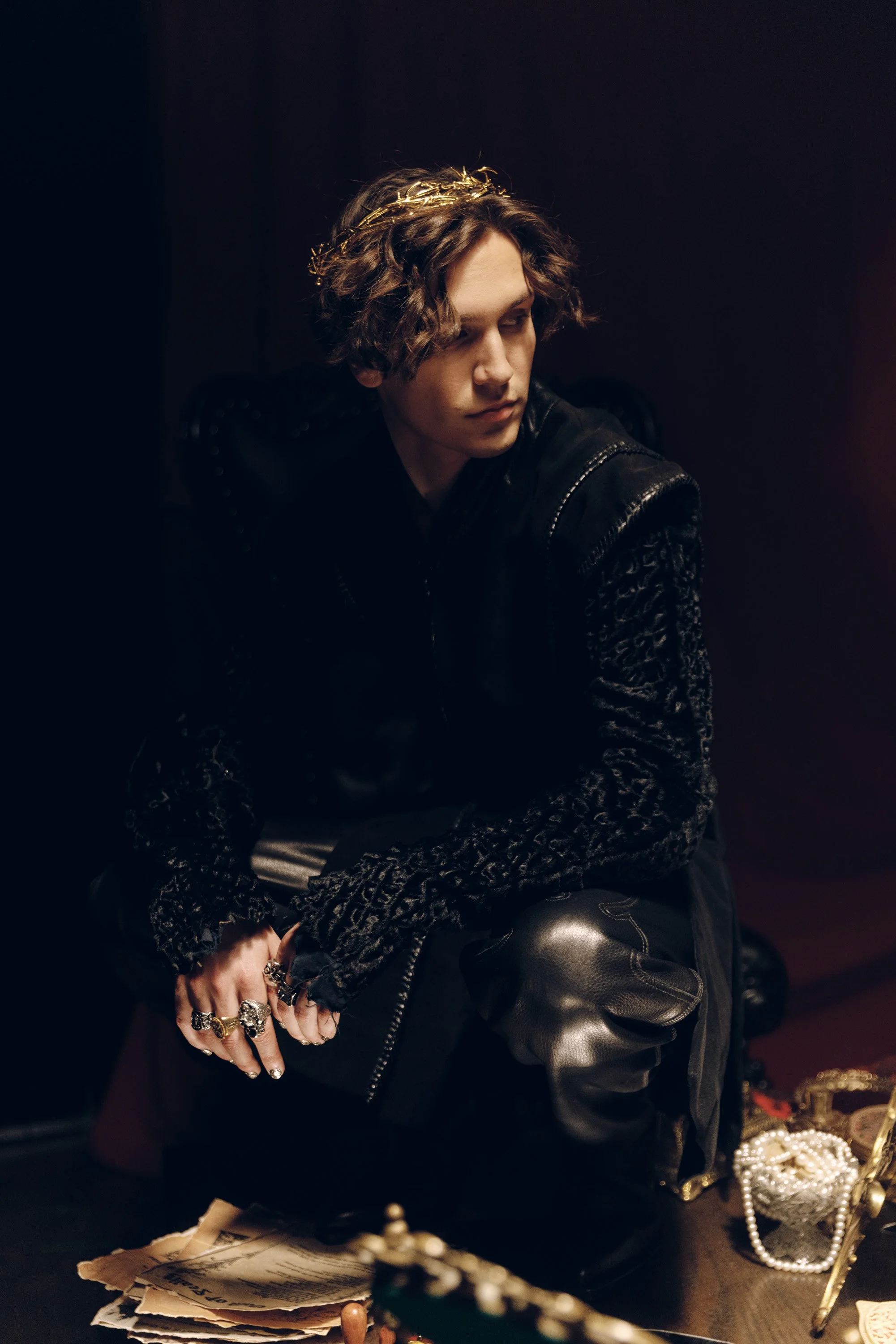 Young person with curly hair wearing a golden crown, black leather jacket, and patterned gloves, sitting in a dimly lit room surrounded by jewelry and vintage items.
