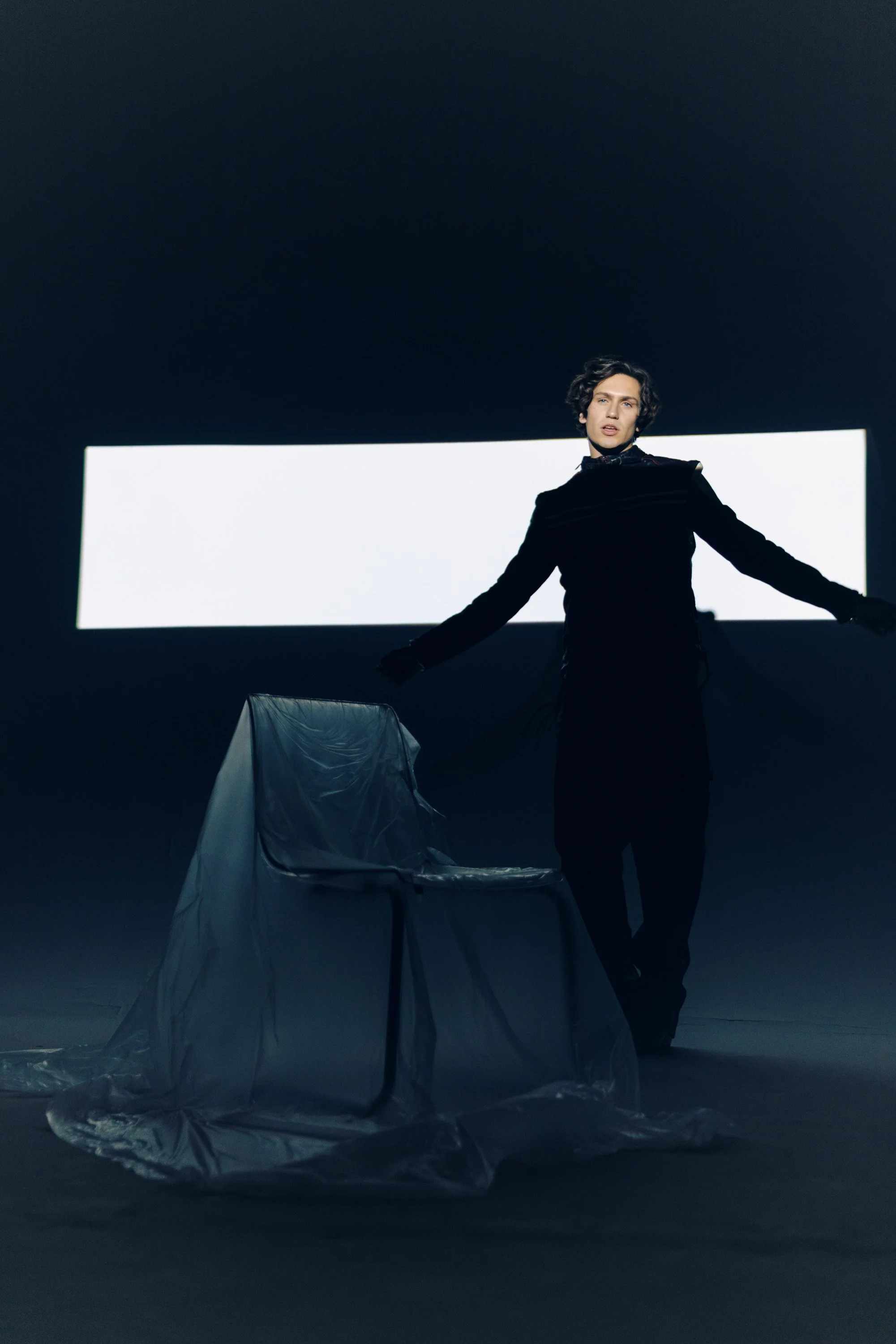 A person with short curly hair dressed in black stands in a dark room with a large white rectangular background. There is a transparent chair and a draped fabric in the foreground. The person appears to be performing or posing, with one arm extended 