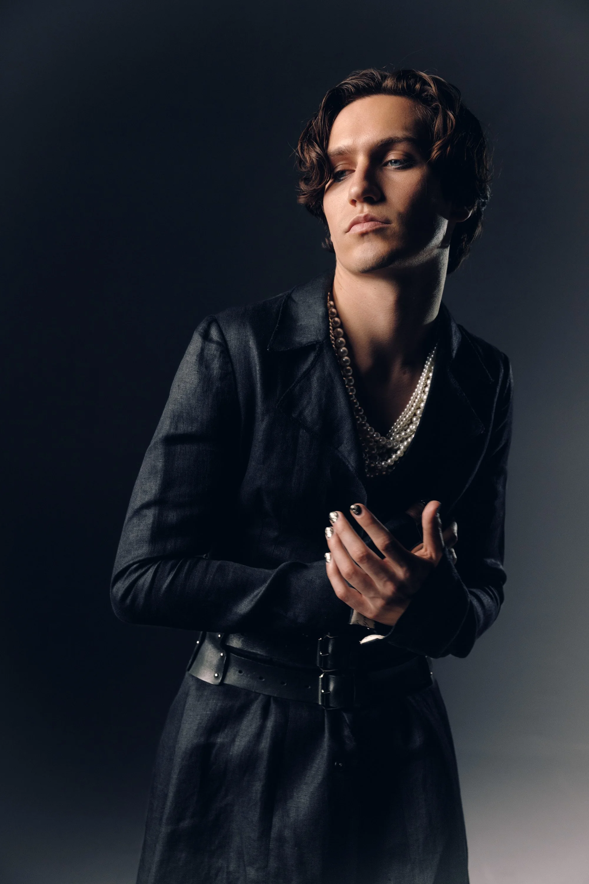 A person with short curly hair and light skin wearing a black leather jacket and multiple pearl necklaces, posing against a dark gray background.