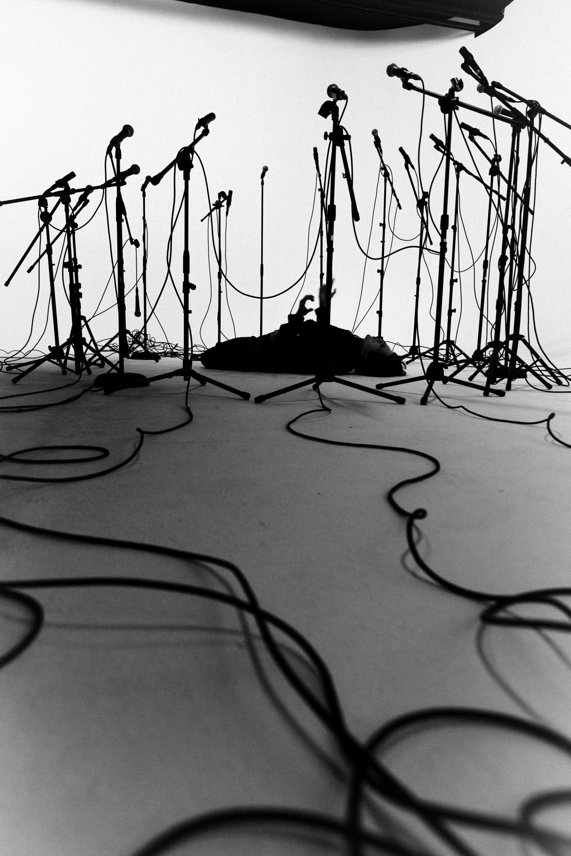 A person lying on the floor surrounded by numerous microphone stands and cables in a studio setting.