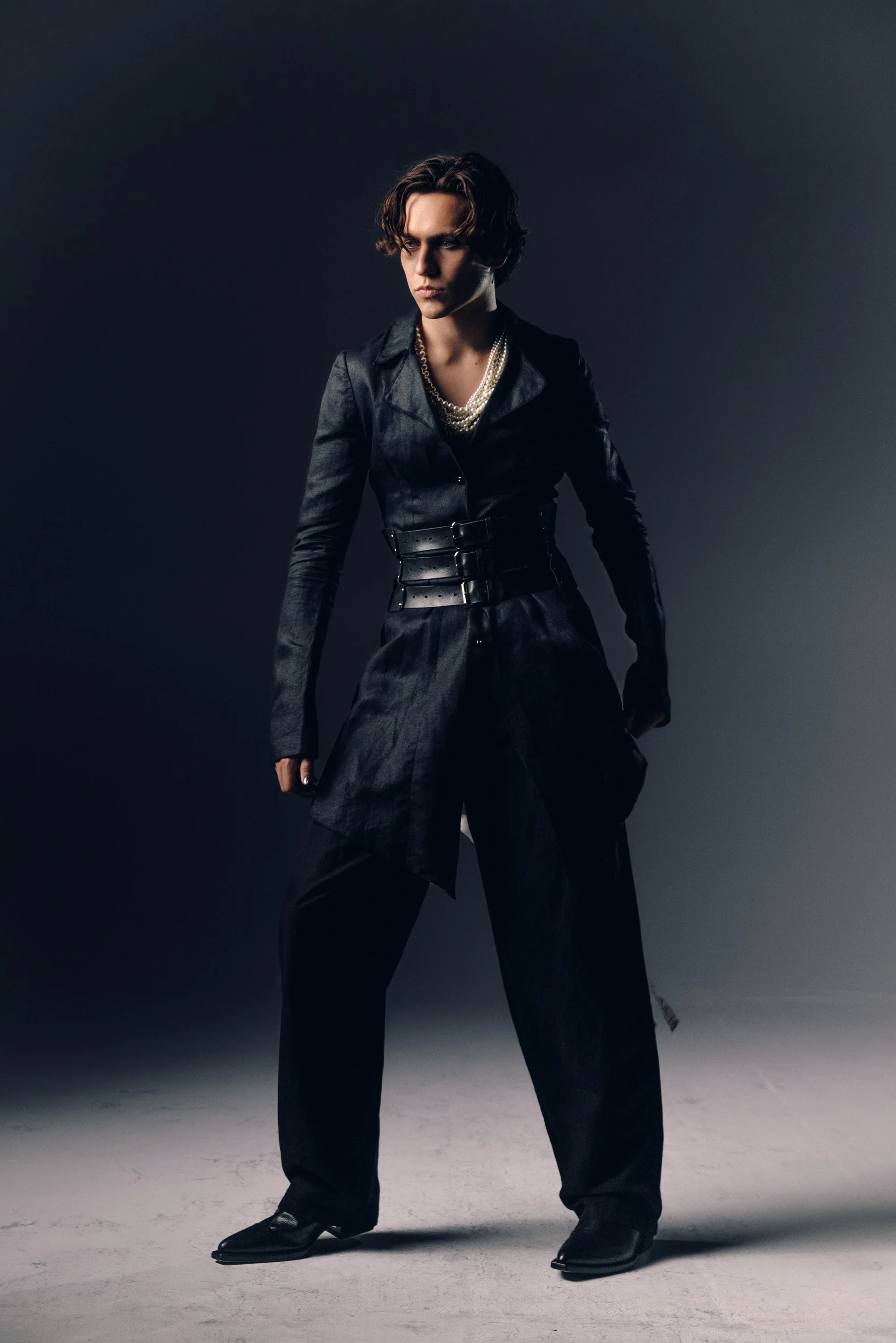 A person with light skin and short, curly hair, wearing a black satin blazer, black belt with large buckle, multiple pearl necklaces, black wide-leg pants, and black shoes, standing on a plain gray floor against a dark gradient background.