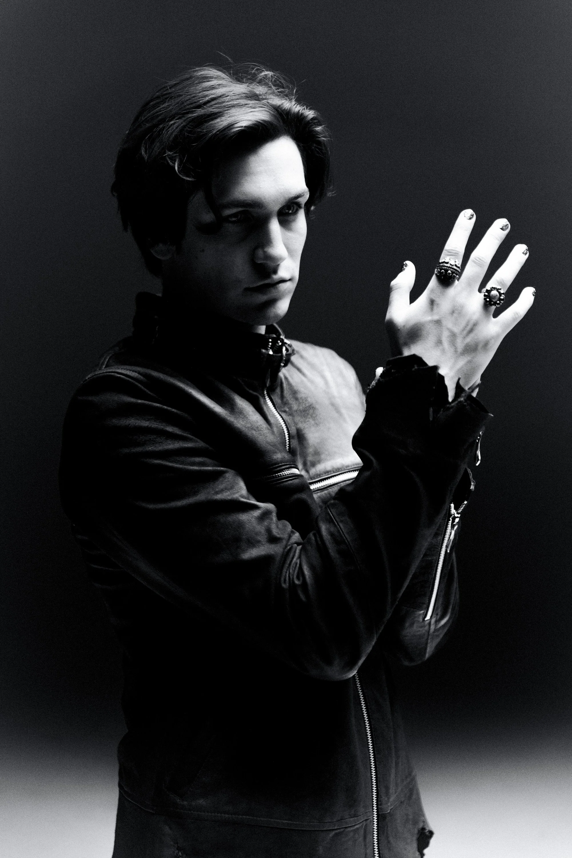 Black and white photo of a person with short wavy hair, wearing a leather jacket, looking at the camera with a serious expression, their hand raised showing multiple rings.