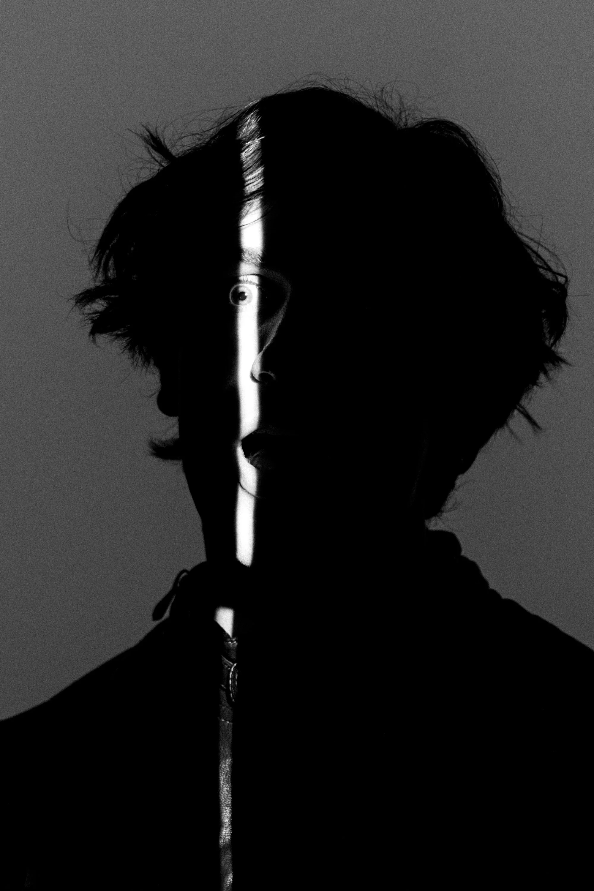 Black and white photo of a person's profile with half of the face in shadow. A vertical light beam creates a stark contrast on the face, highlighting the eye and nose. The person's hair is tousled, and they are wearing a hoodie.