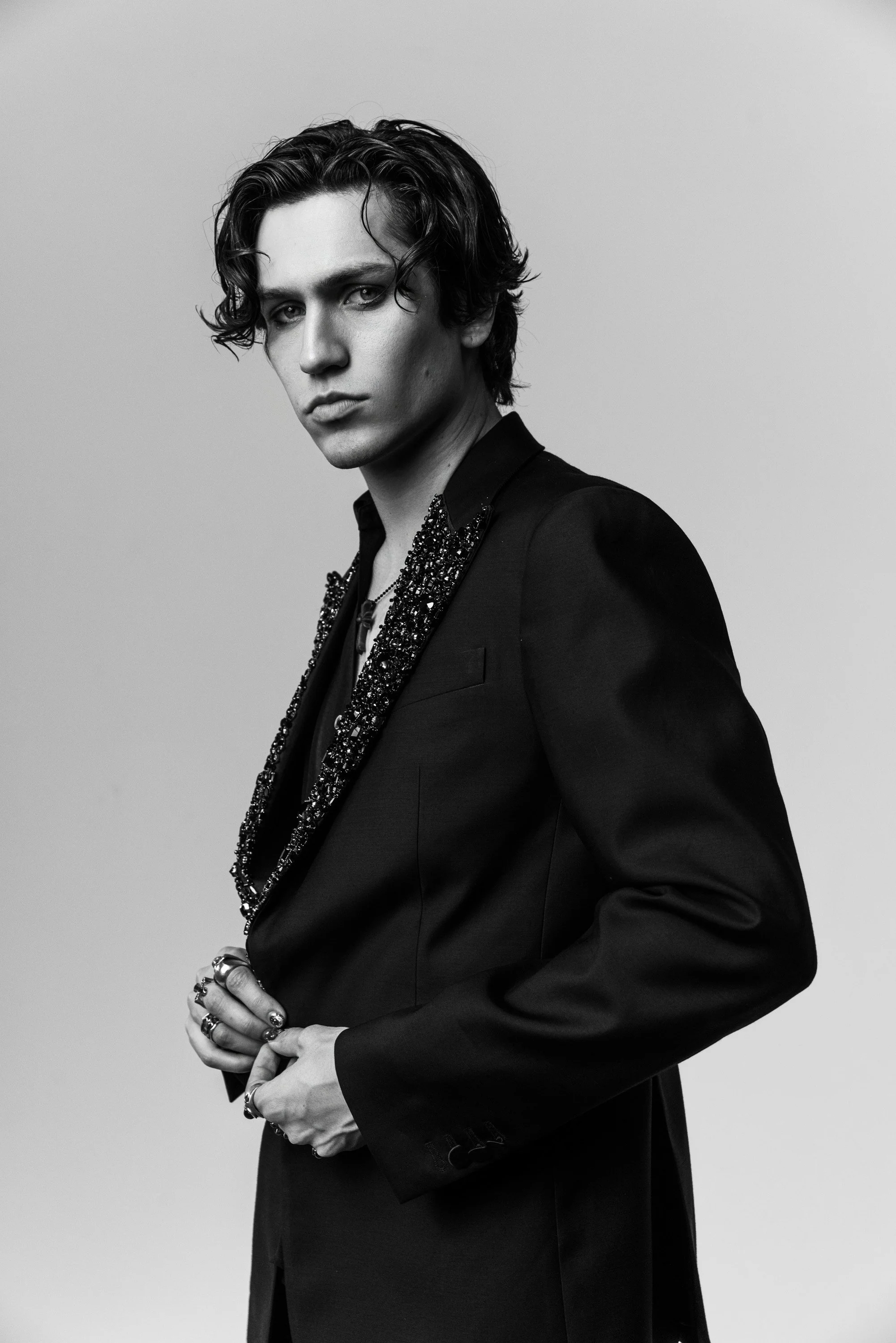 Black and white portrait of a young man with wavy hair, wearing a tailored jacket with embellished lapel, adorned with rings and jewelry, standing against a plain background.
