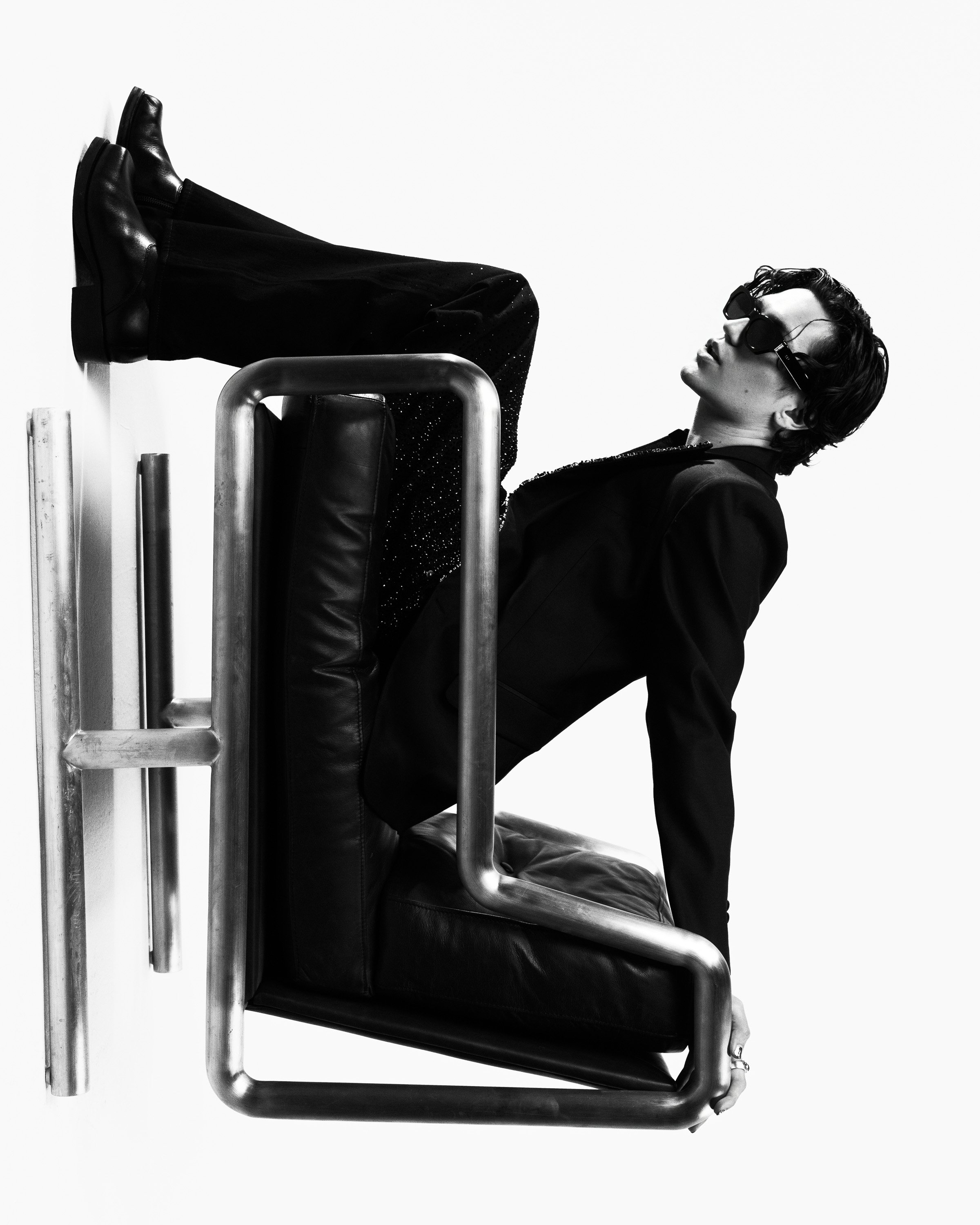 A person with short hair, sunglasses, and stylish clothing sitting on a rolling chair with their legs resting on the seat, using a minimalist, monochromatic setting.