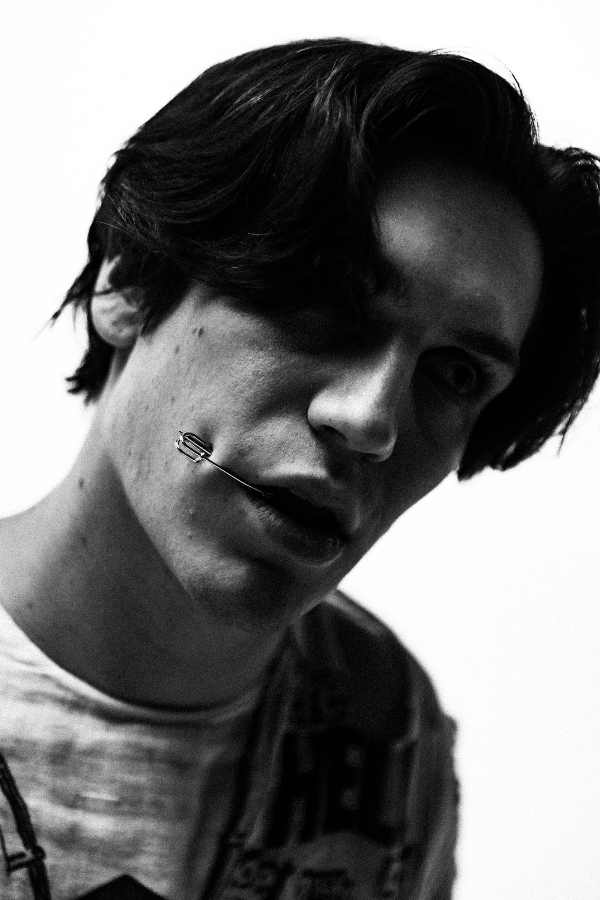 Black and white close-up photo of a person with short dark hair, partially visible face, and a piercing on the lower lip, wearing a dark jacket and a hoodie.