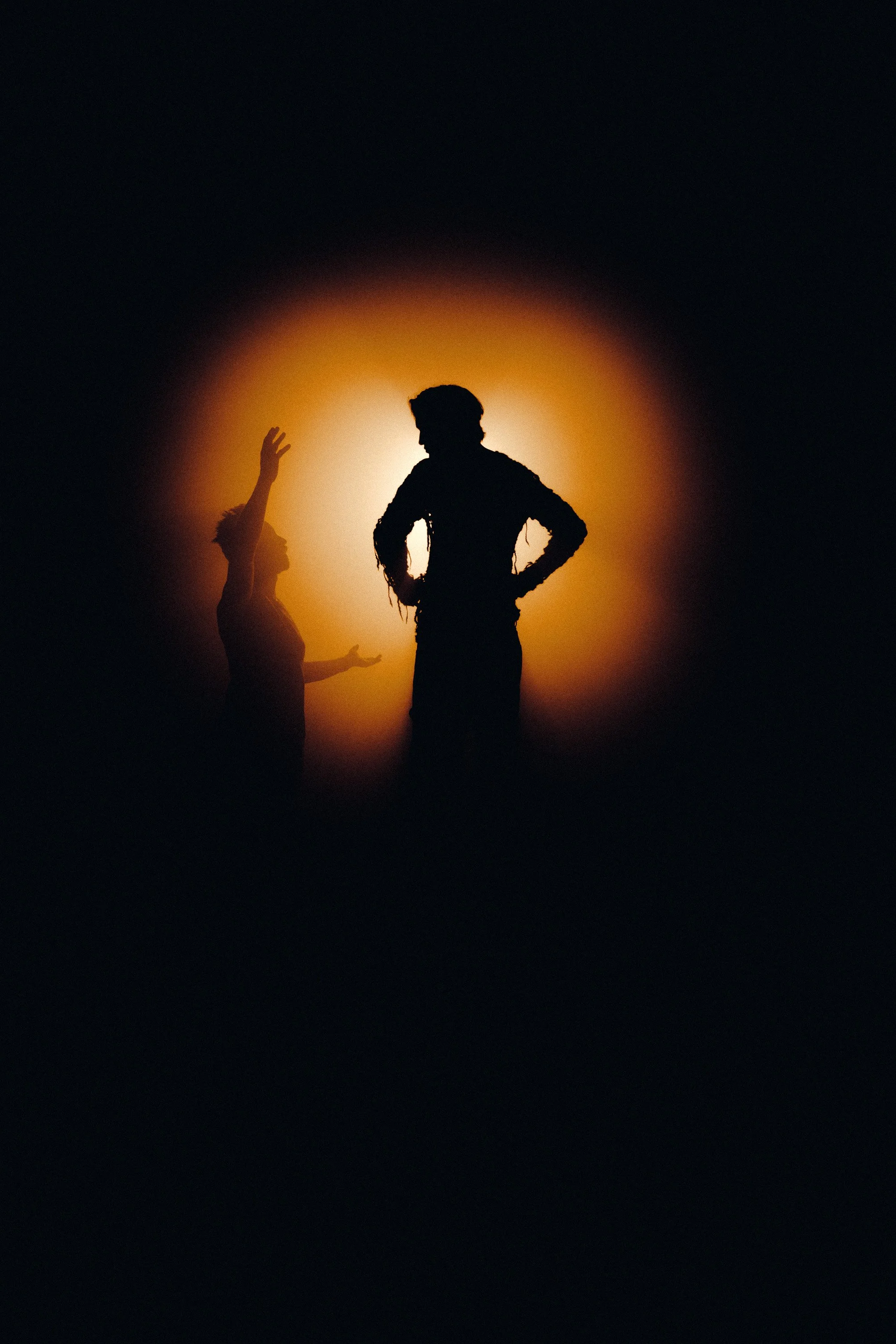 Silhouettes of two people, one with hands on hips and the other reaching up, standing against a bright, warm backlight in a dark setting.