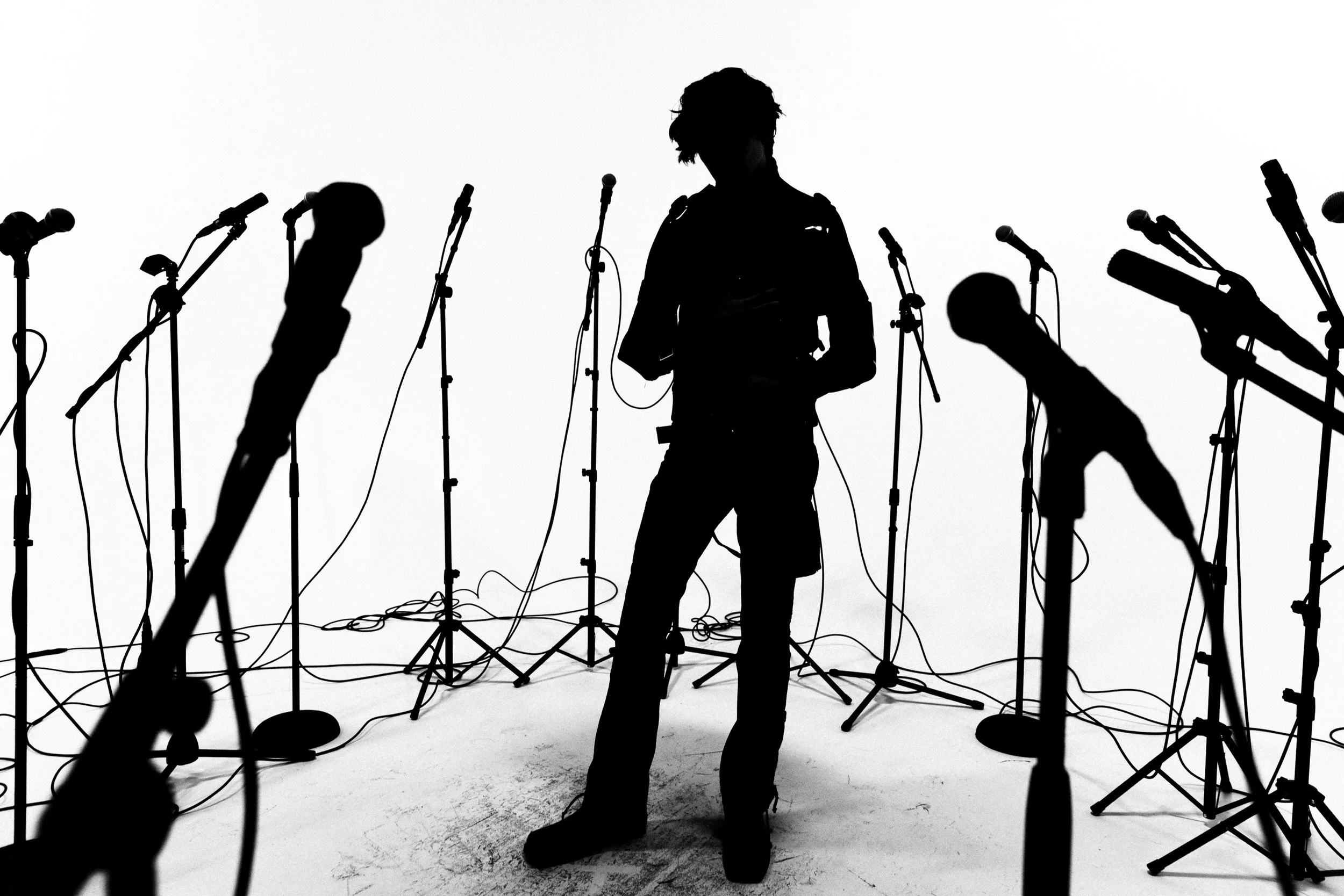 Silhouette of a person standing among multiple microphone stands in a studio setting, with a white background.