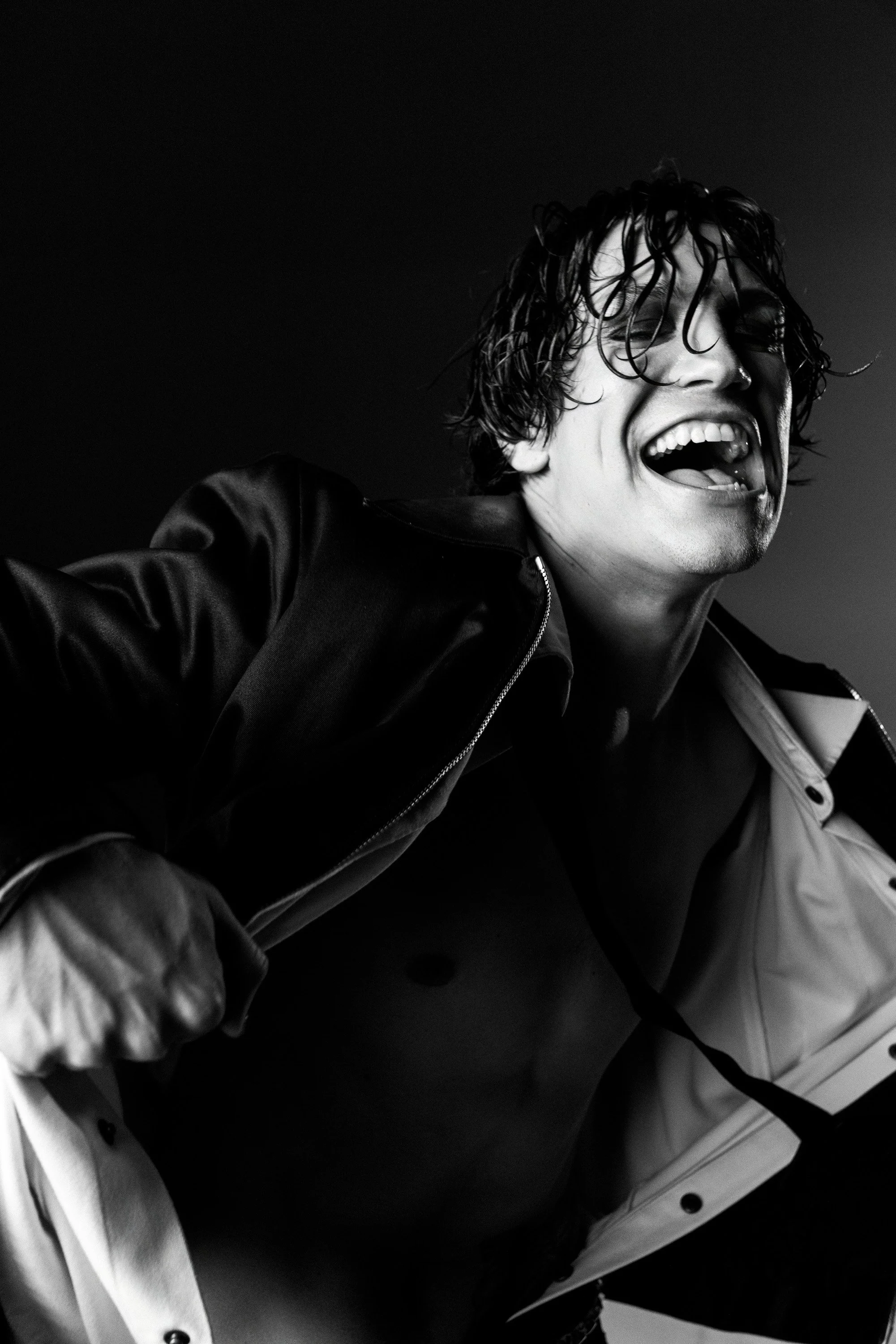 Black and white photo of a person with curly hair, smiling widely with mouth open, wearing a shiny jacket, sitting on a chair.