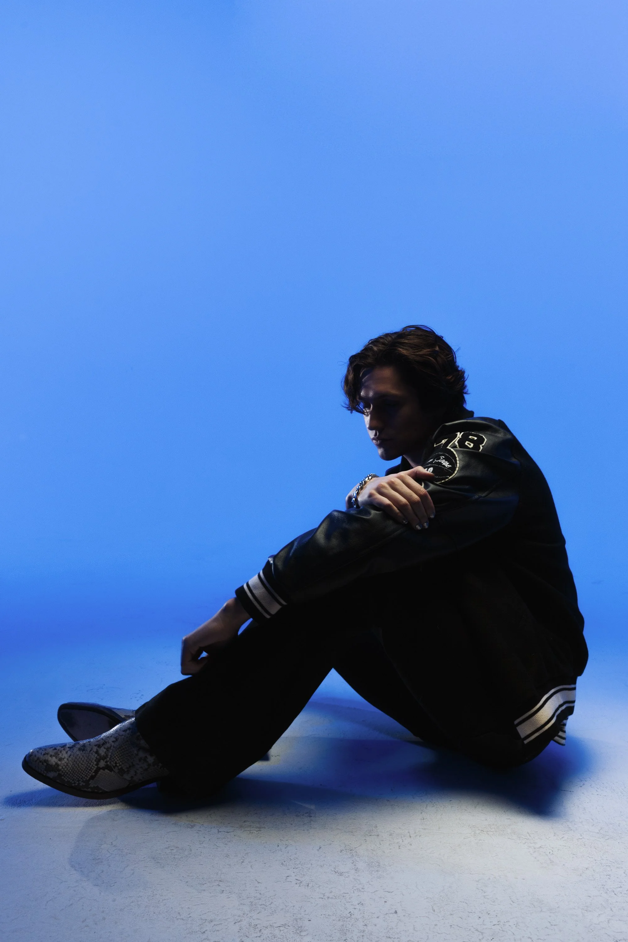 A young man with wavy hair sitting on the floor against a blue background, dressed in a black letterman jacket with white and black striped cuffs, black pants, and snakeskin patterned shoes, looking downward with a serious expression.