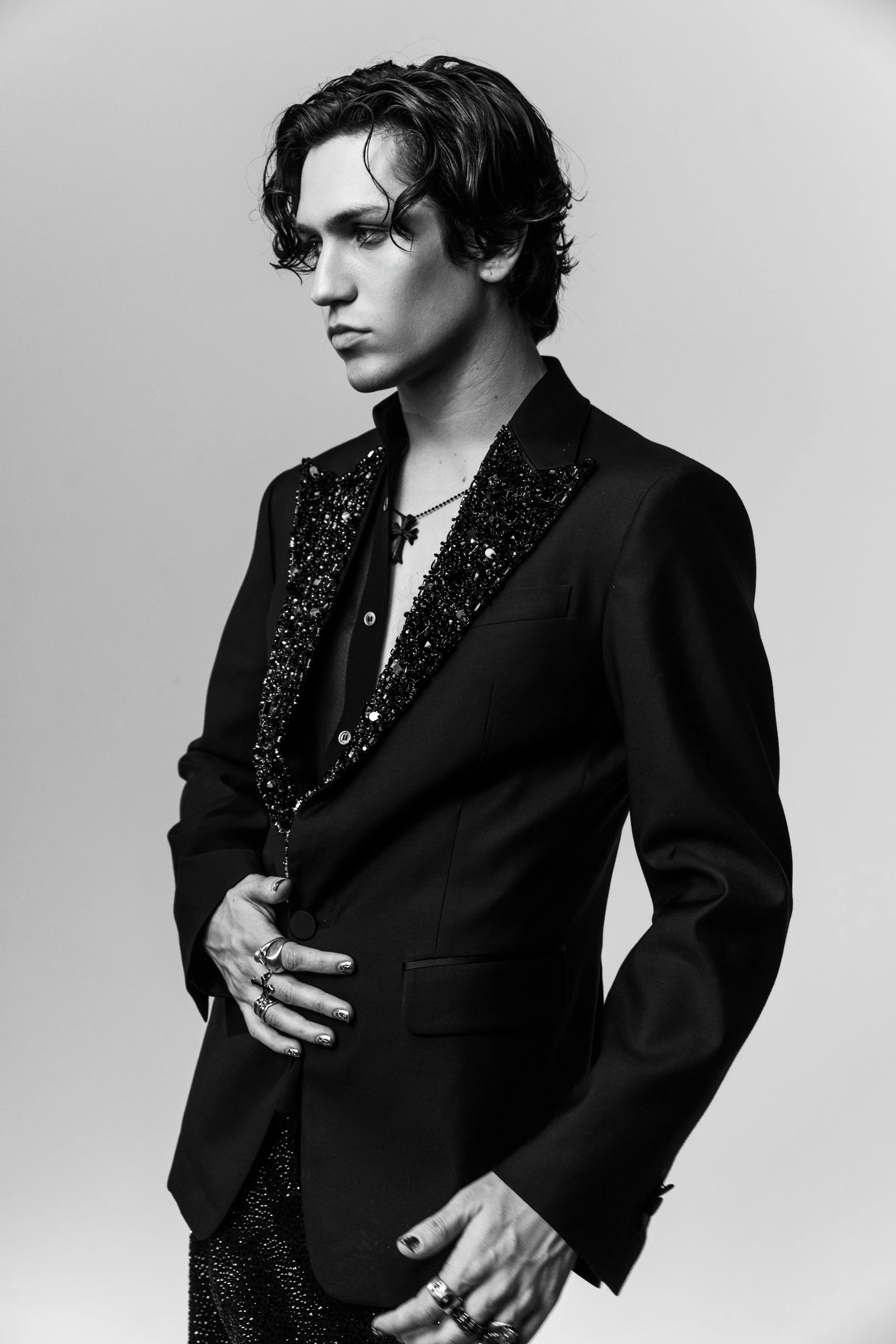 A black-and-white portrait of a young man with wavy dark hair, dressed in a black tailored jacket with beaded embellishments on the lapels, wearing multiple rings and a necklace, with a contemplative expression, against a plain background.