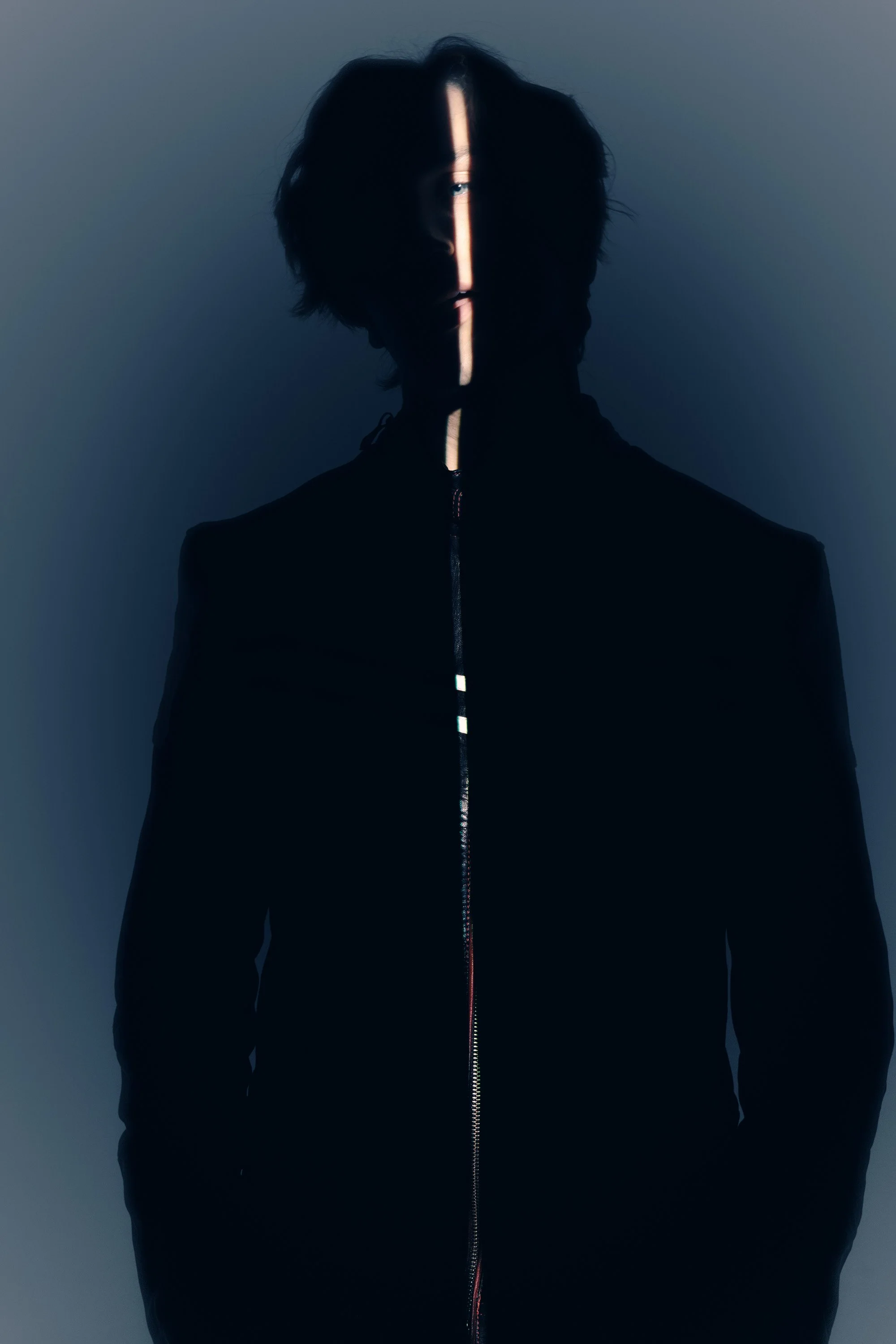 A person with dark, wavy hair is standing against a plain background. The image is dark, with a narrow vertical light strip illuminating part of their face and the zipper of their jacket.