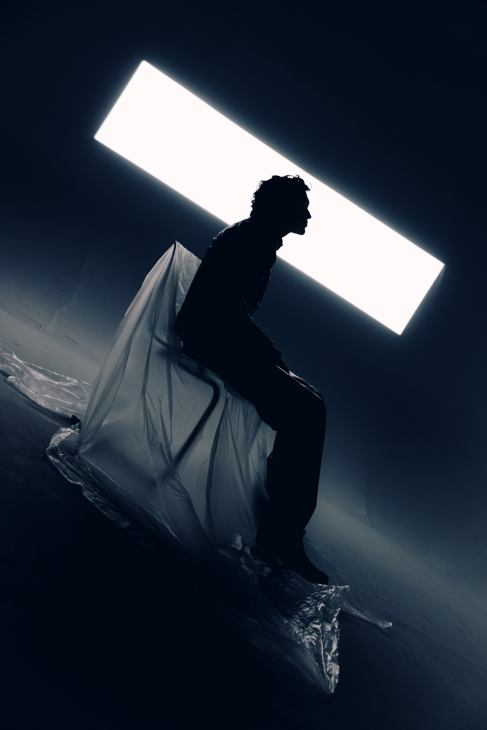 Silhouette of a person sitting on a chair with a large illuminated rectangular light source above, with reflective material on the floor.