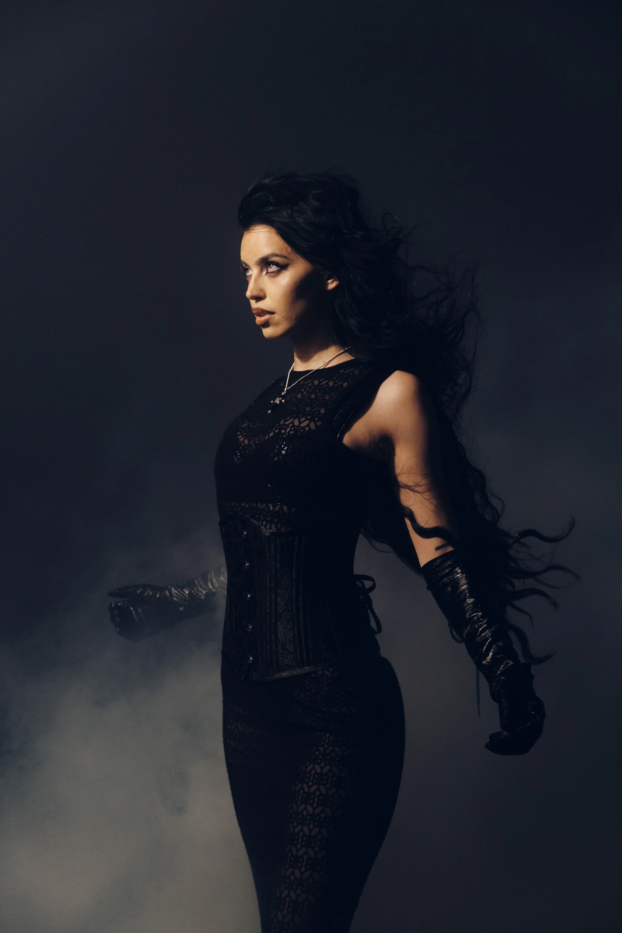 A woman with long black hair flowing in the wind, dressed in a black, lace and leather outfit, posing against a dark, smoky background.