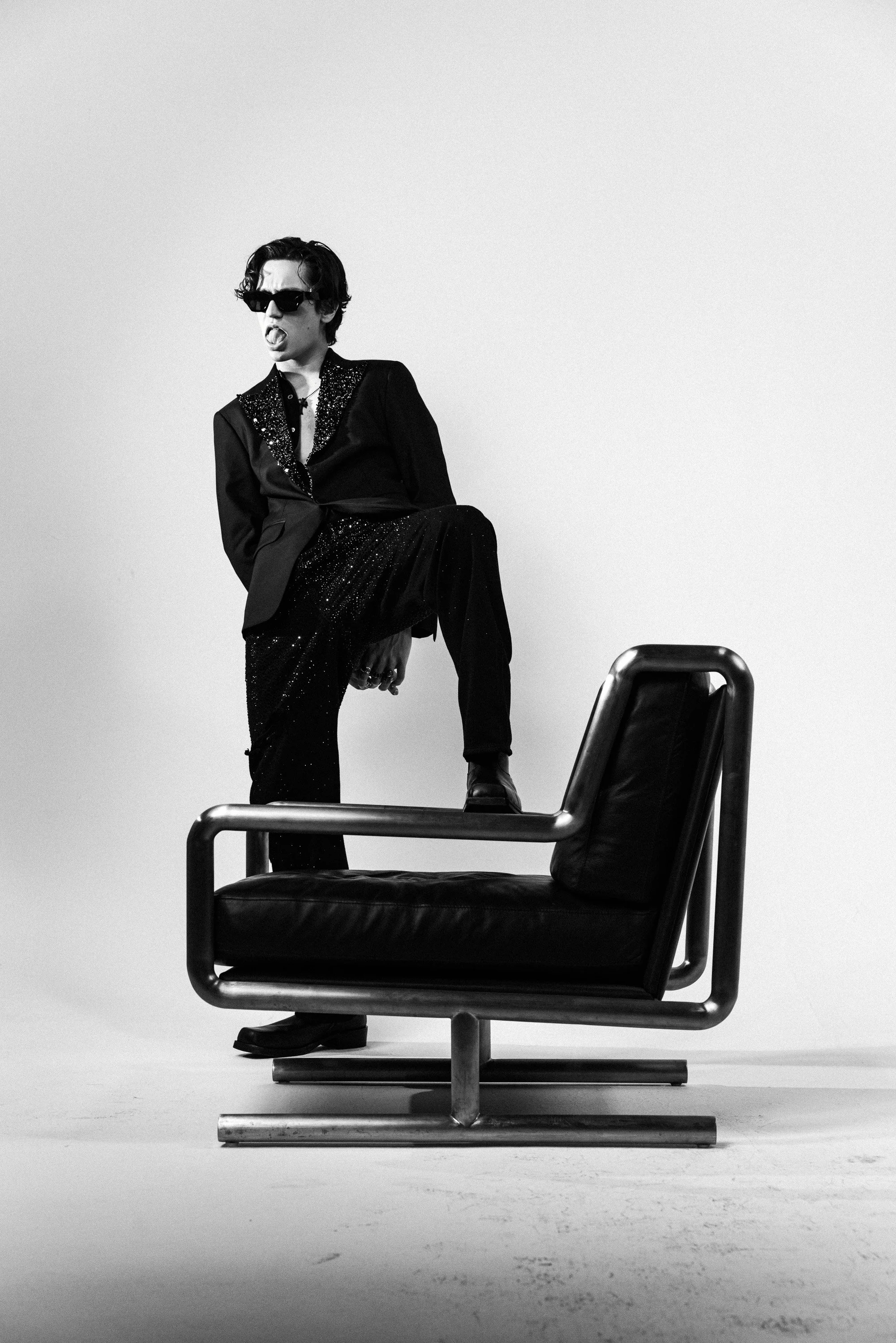 A person wearing sunglasses and a sparkly jacket, sitting with one leg propped on a chair, in a minimalist setting with plain walls and a modern office chair.