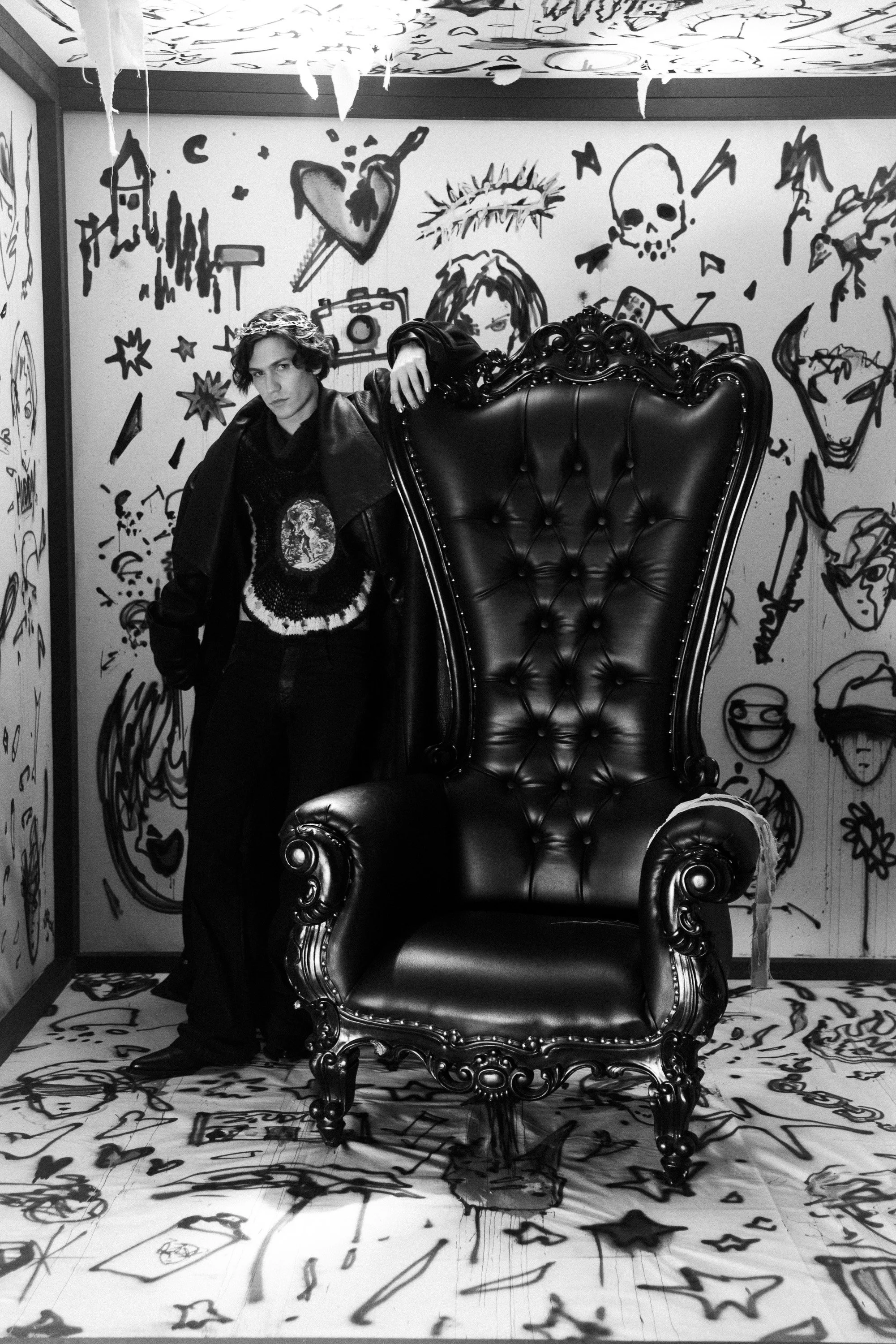 A young man with dark, wavy hair standing in front of a large, ornate black leather throne with intricate detailing. The background, floor, and ceiling are decorated with various black graffiti-style drawings and sketches, including skulls, hearts, f
