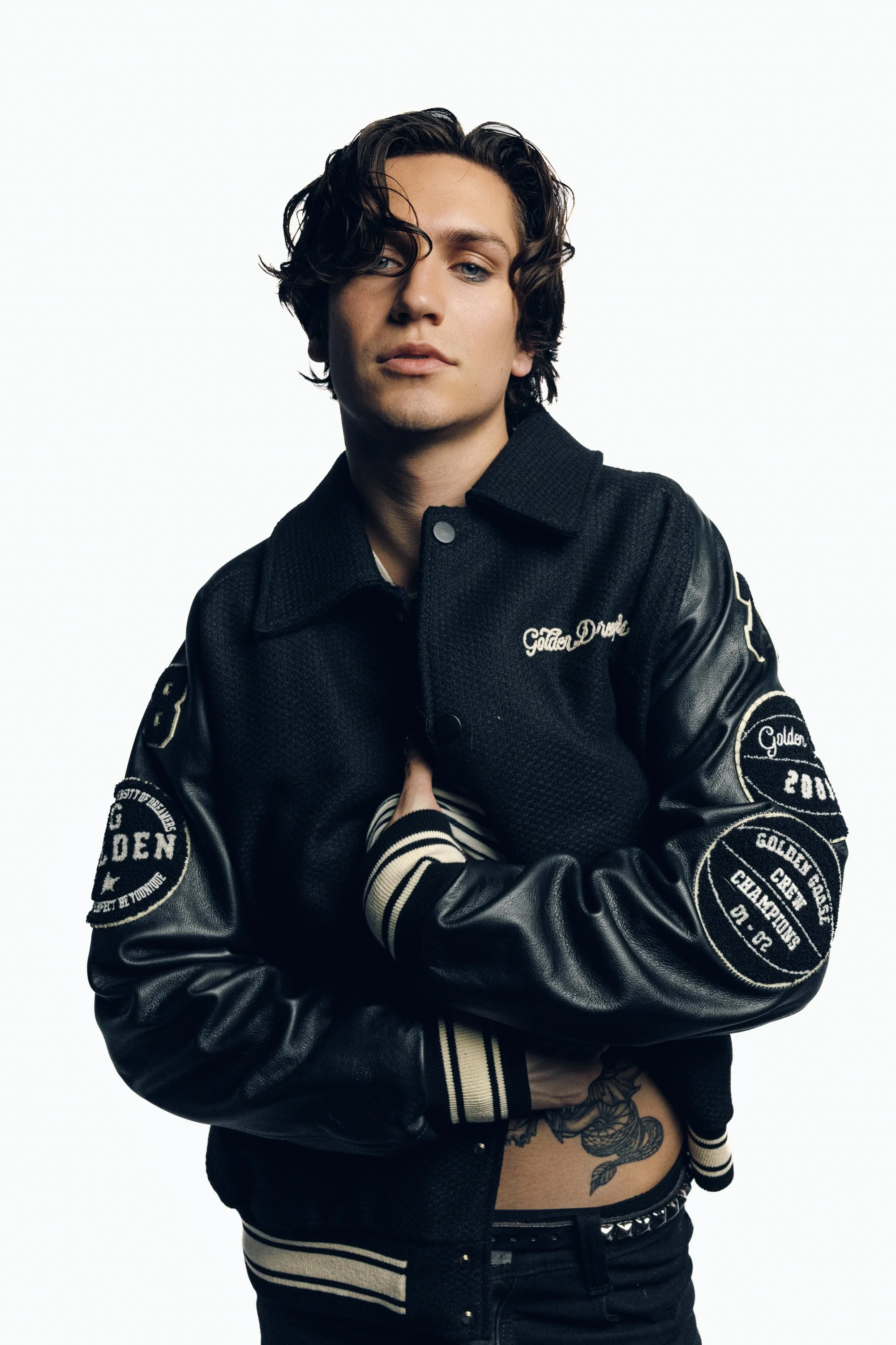 A young man with wavy dark hair and light skin poses against a plain white background. He's wearing a black varsity jacket with patches on the sleeves and the words 'Golden Drive' embroidered on the chest. His left hand is resting on his chest, and h