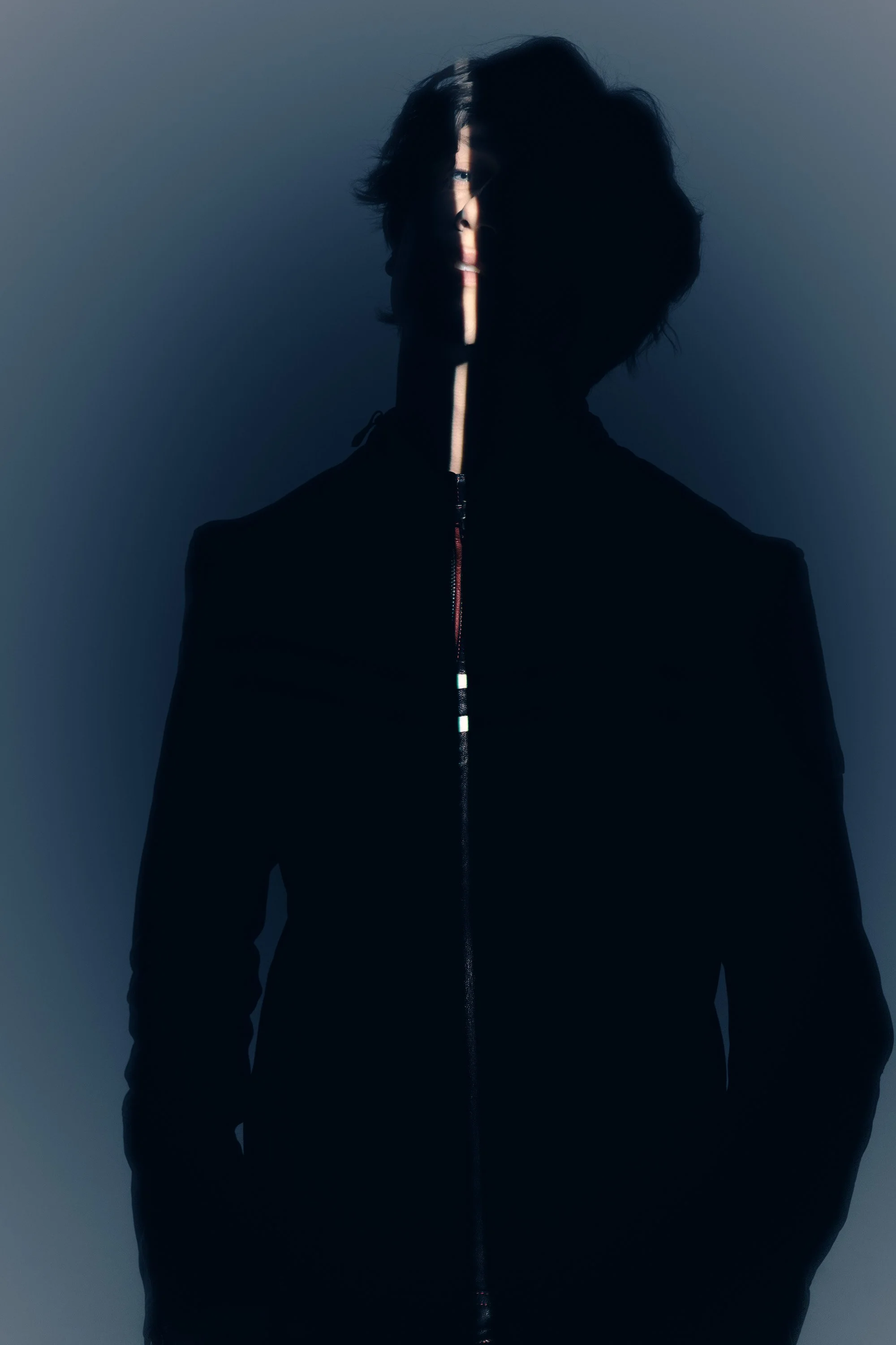 Person with tousled hair wearing a black jacket, standing against a plain background, with a shadow of a vertical light strip partially illuminating their face.