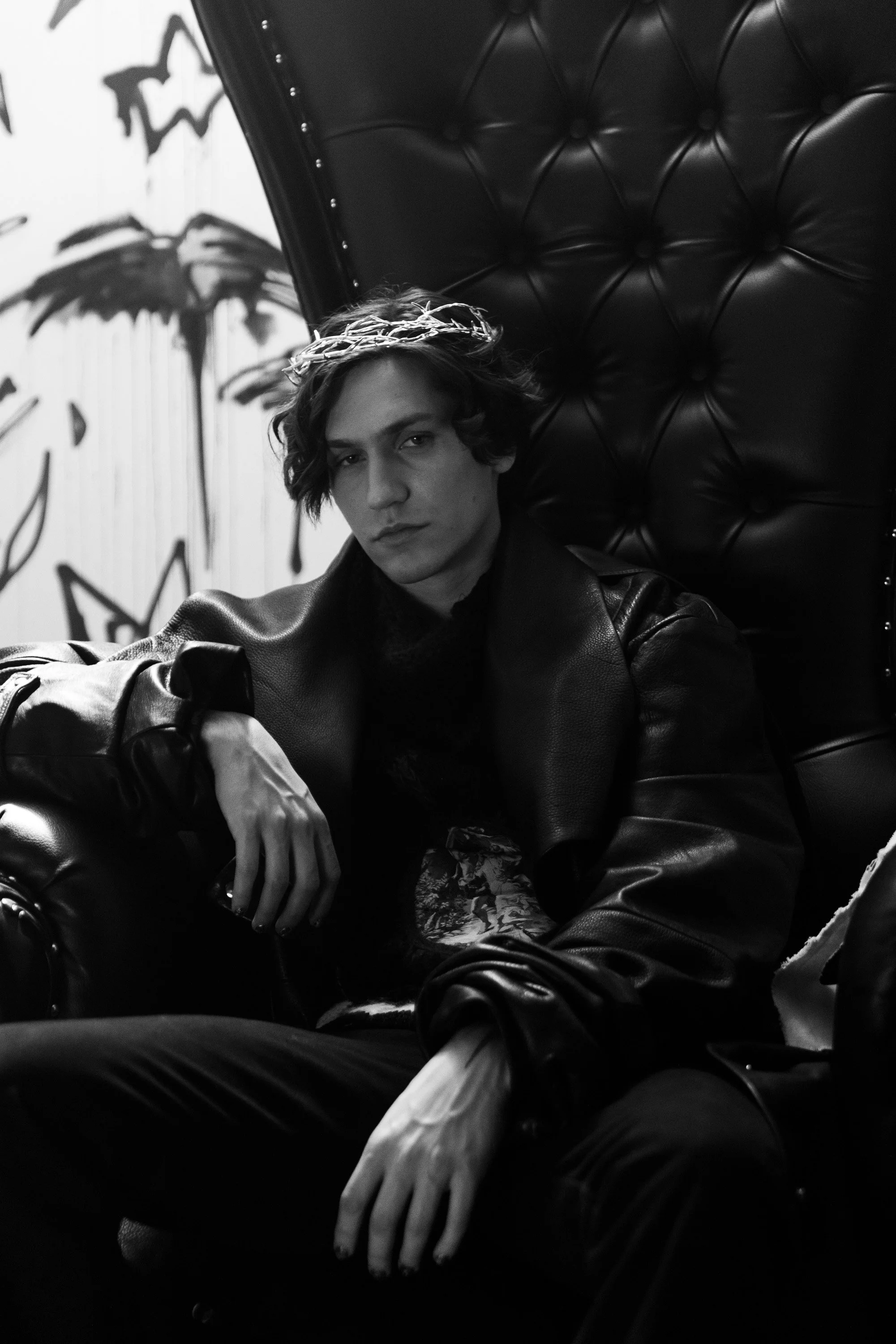 Black and white photo of a young man with dark hair wearing a leather jacket, sitting on a leather armchair with a tufted backrest and a crown-shaped headband, in front of a wall with graffiti artwork.