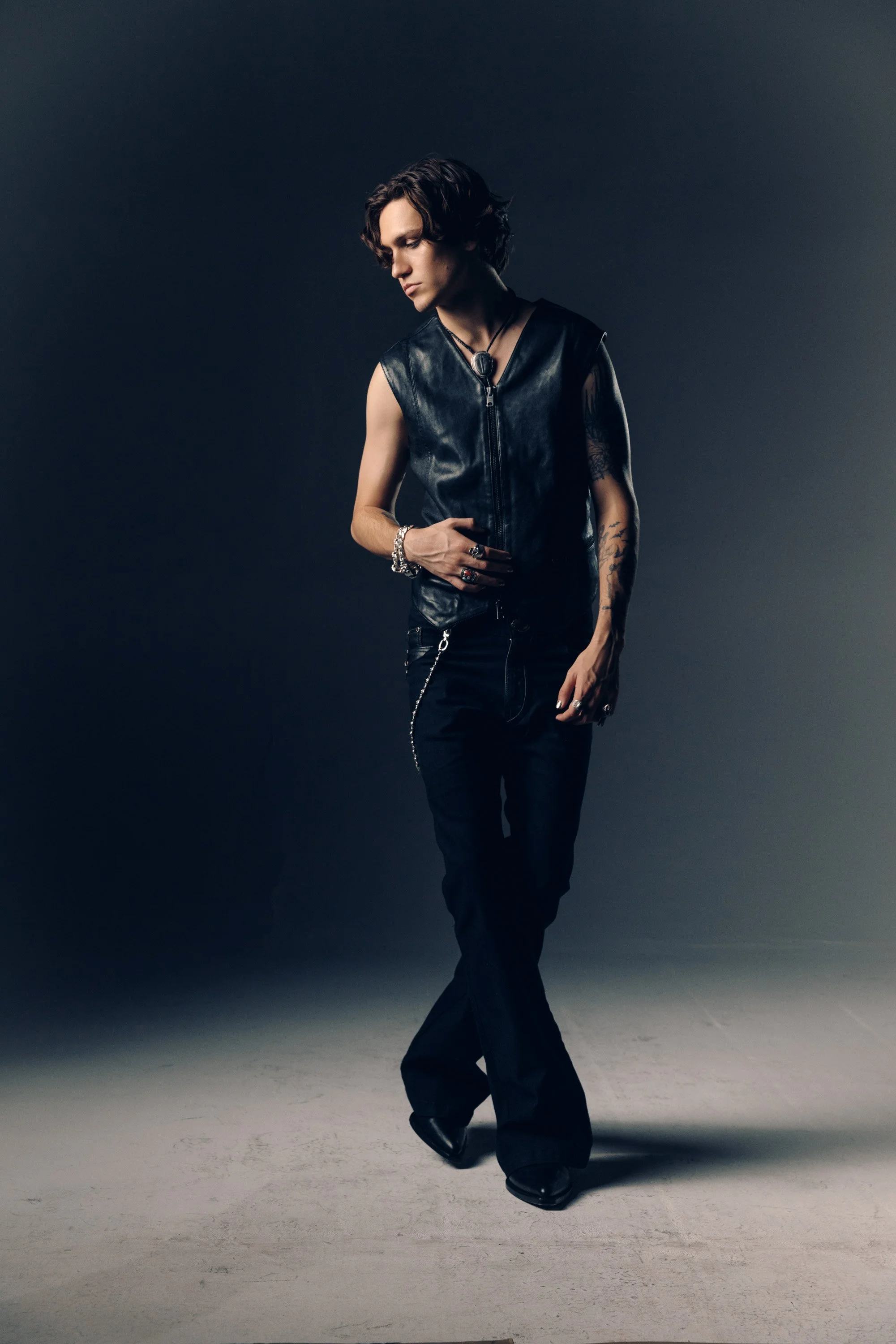 A person with short wavy hair and tattoos, dressed in a sleeveless leather vest and black pants, stands against a dark gradient background. They are wearing jewelry, including rings, bracelets, and a necklace, and look downward with a contemplative e