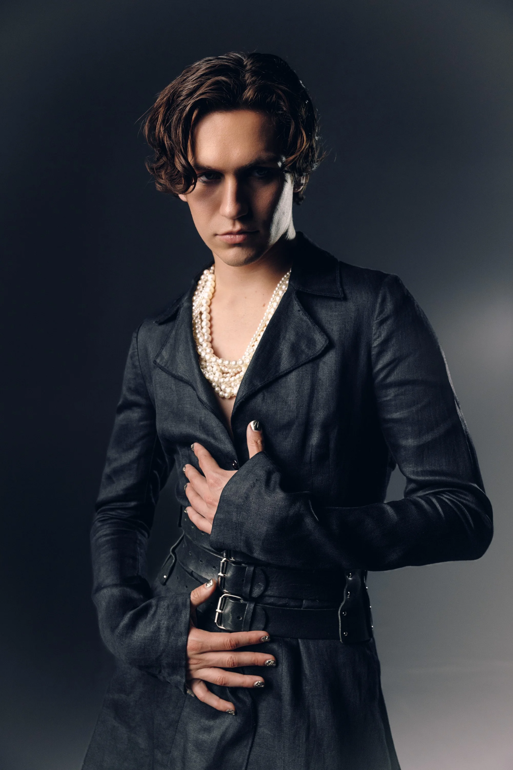 A person with short, wavy brown hair wearing a black leather jacket, pearl necklace, and with painted nails. They are posing with one hand on their chest and the other on their waist against a dark background.