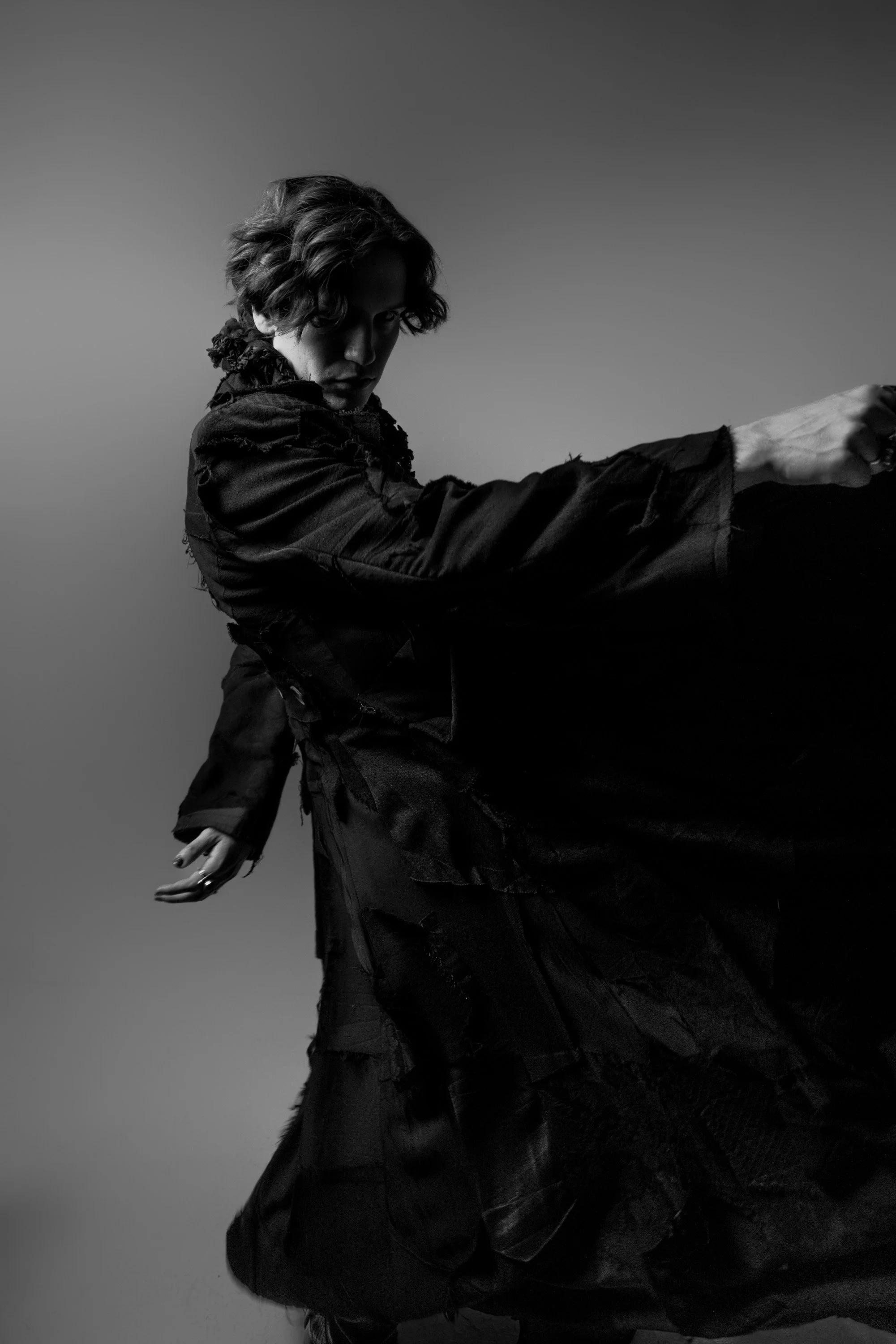 A black and white photo of a person with wavy hair in motion, wearing dark clothing, and striking a dynamic pose against a plain background.