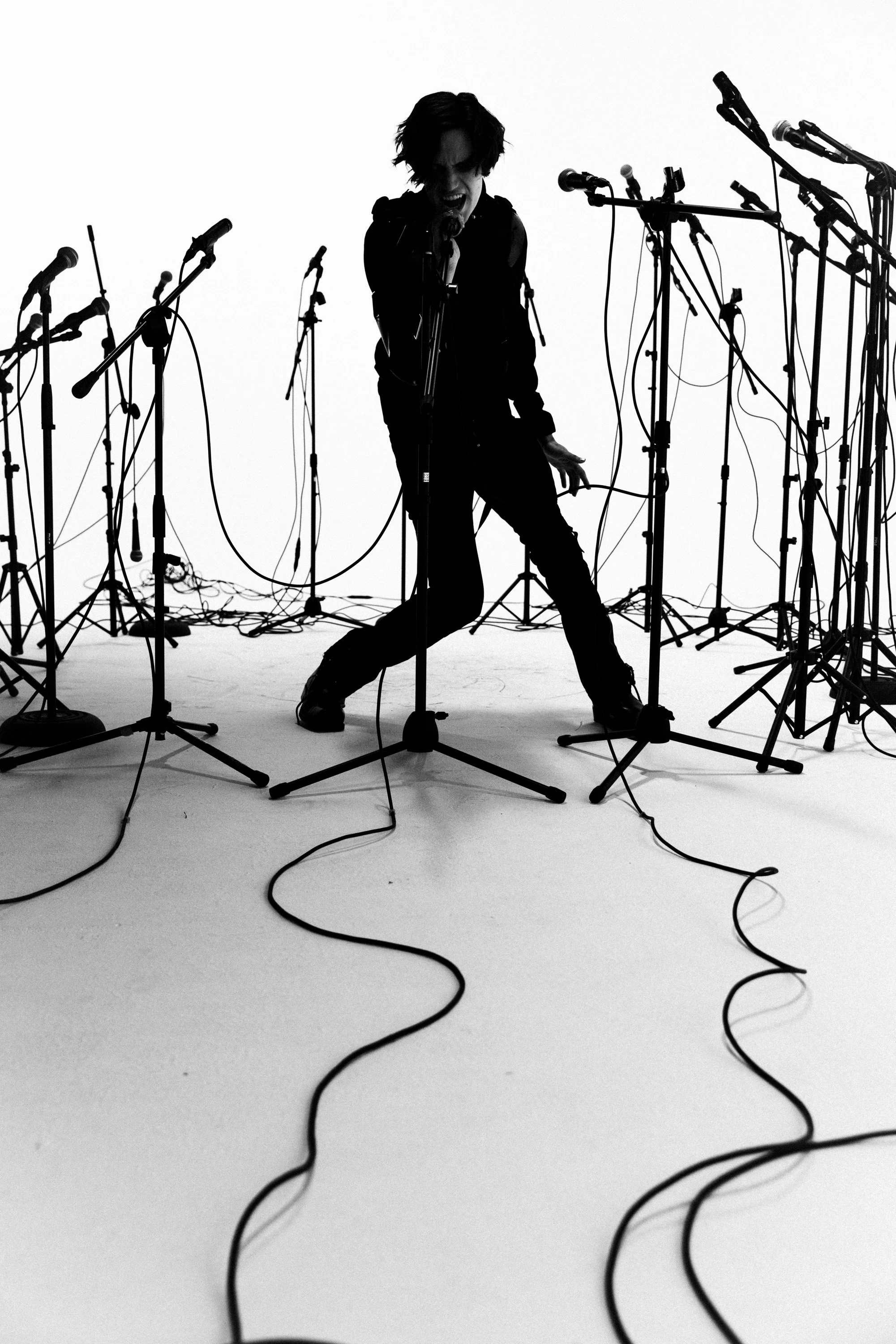 Black and white photo of a woman singing into a microphone, surrounded by multiple microphone stands and wires, in a studio or performance space.