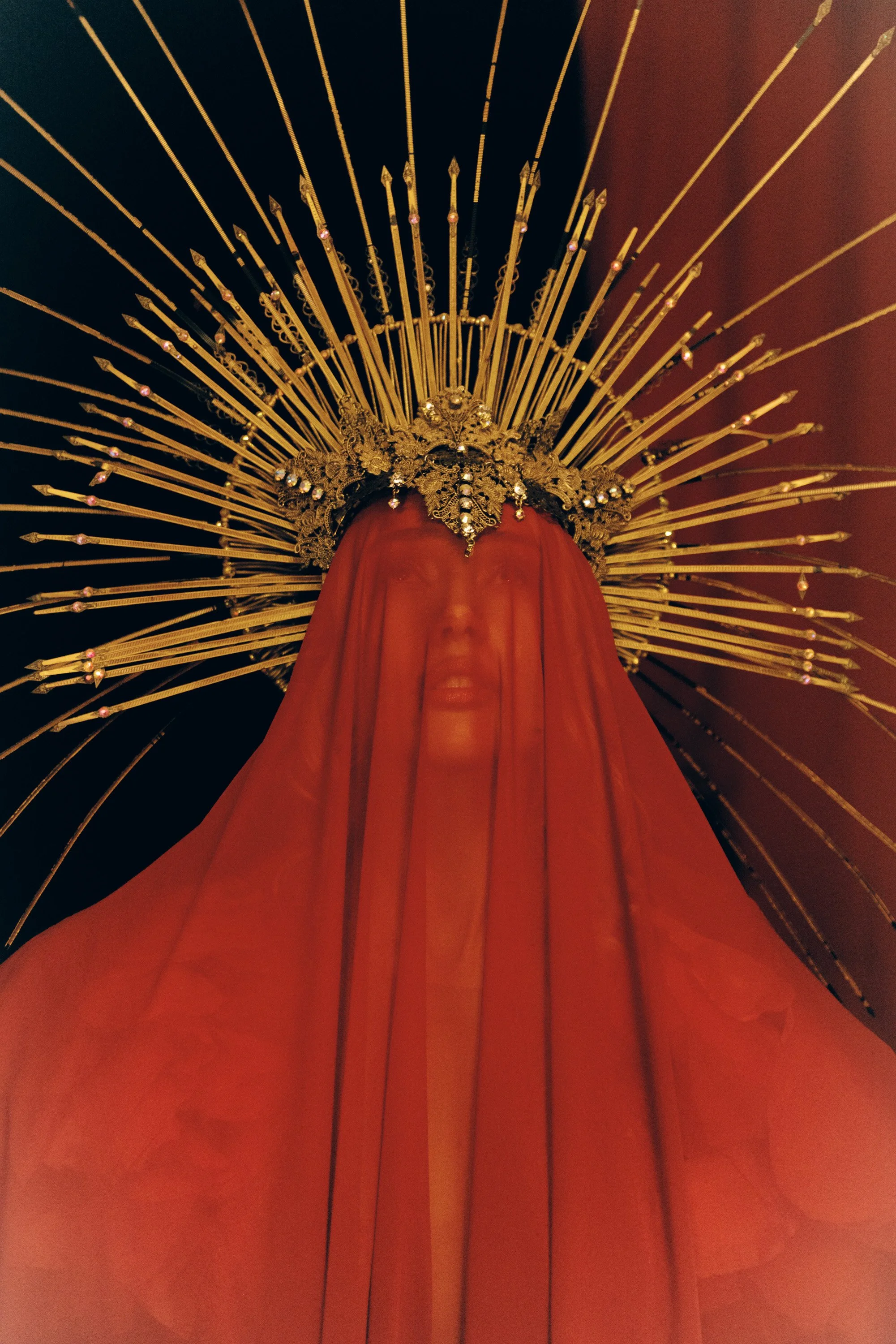 Close-up of a religious statue with a golden ornate crown and a red cloth covering its face, surrounded by a halo of long golden rays.