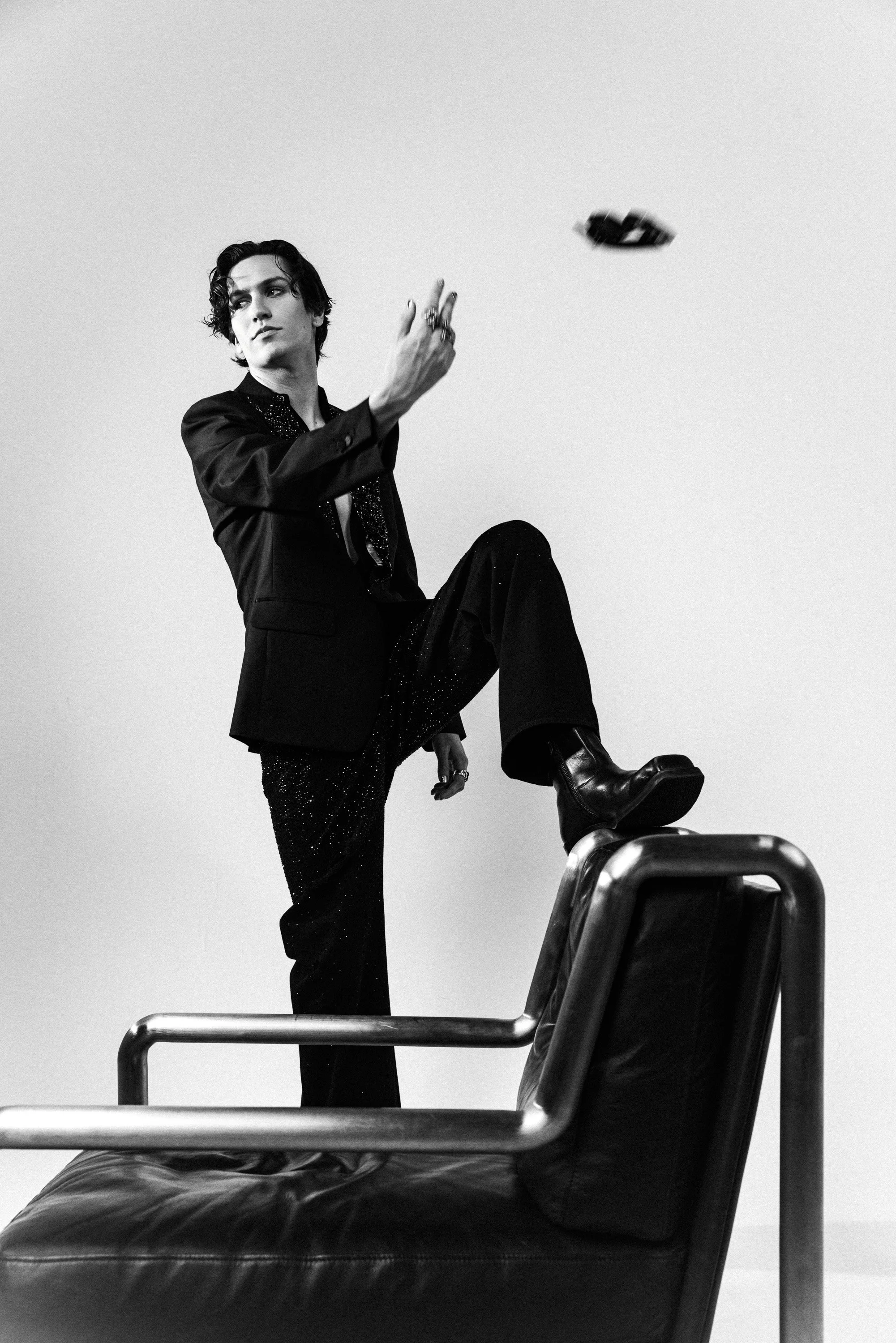 A black and white photo of a young man with dark, wavy hair, wearing a black blazer and pants with a sparkly texture. He is sitting on a chair, with one leg raised and resting on a metal railing, while his hand is mid-air, holding a car key or remote