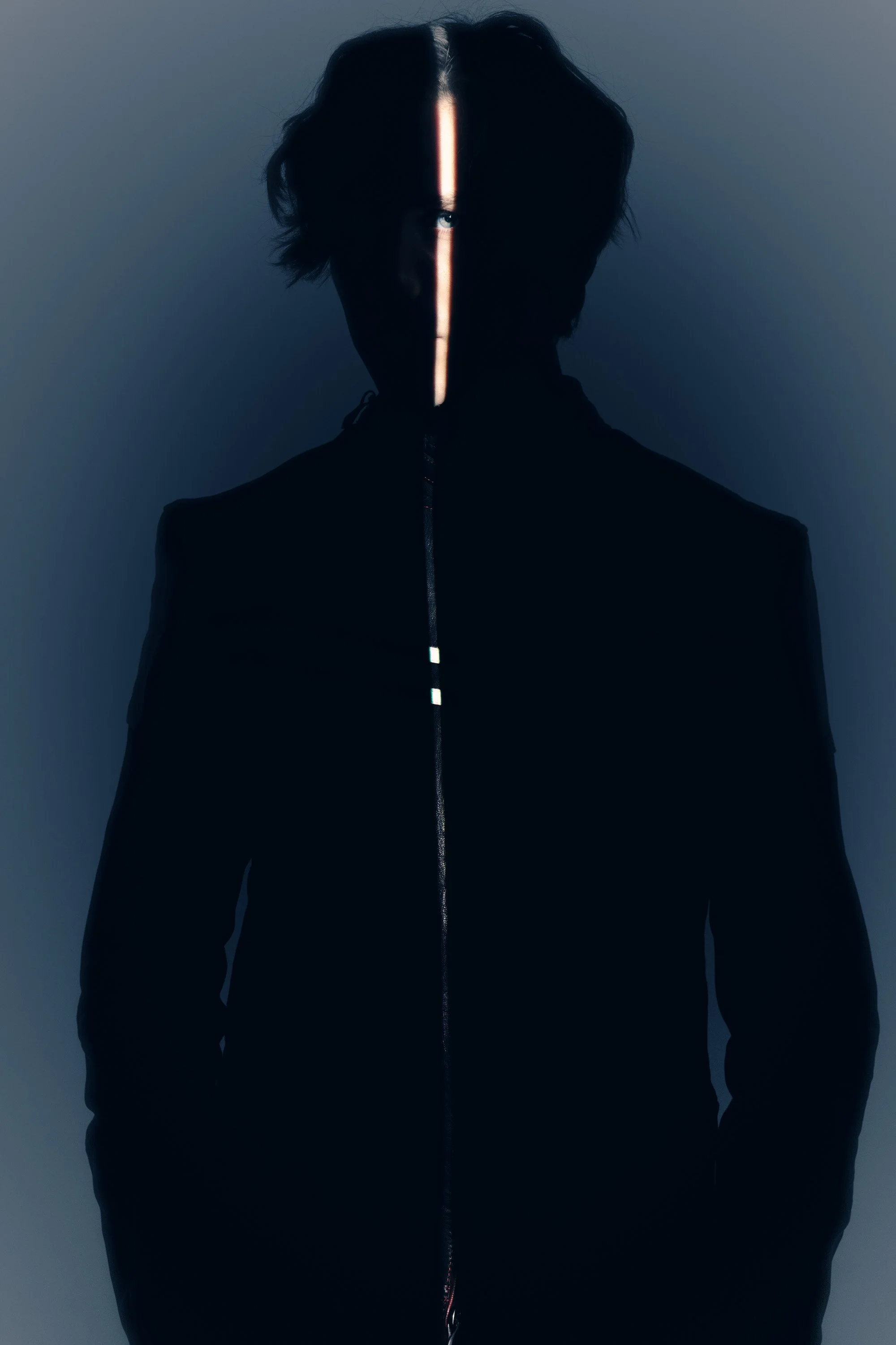 A person with black hair and light eye color, wearing a black jacket with a reflective front zipper, standing against a plain gray background. Their face is partially obscured by a vertical light beam.